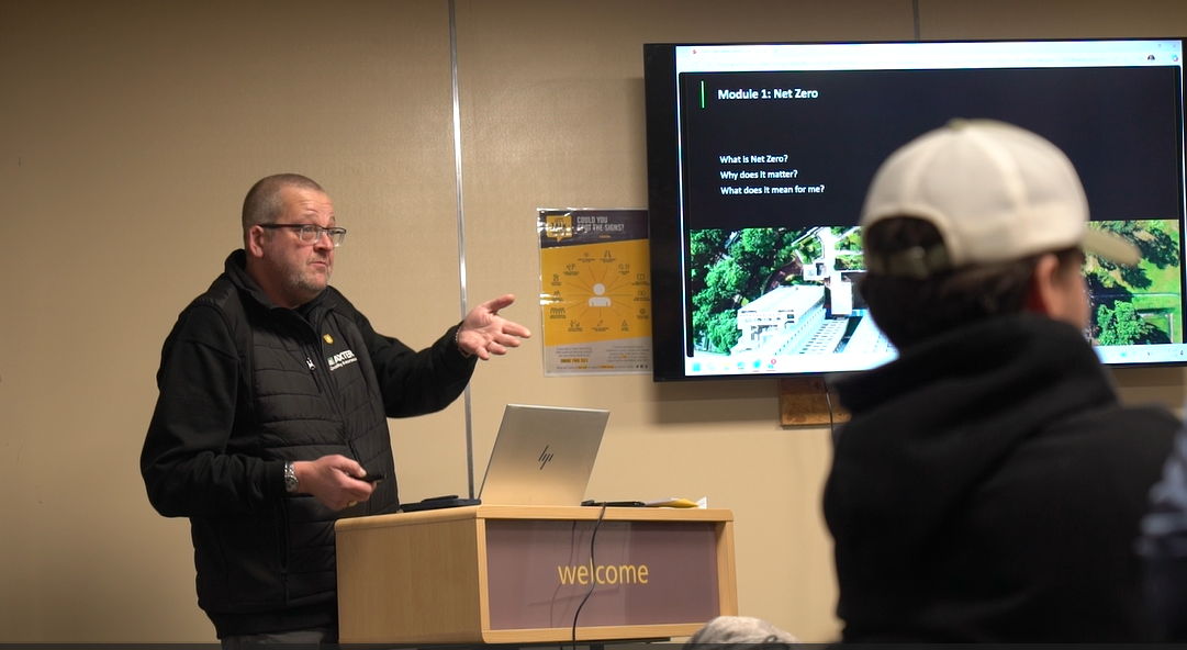 Man in dark jacket presenting to audience using laptop and wall-mounted screen displaying green forest imagery in beige conference room.