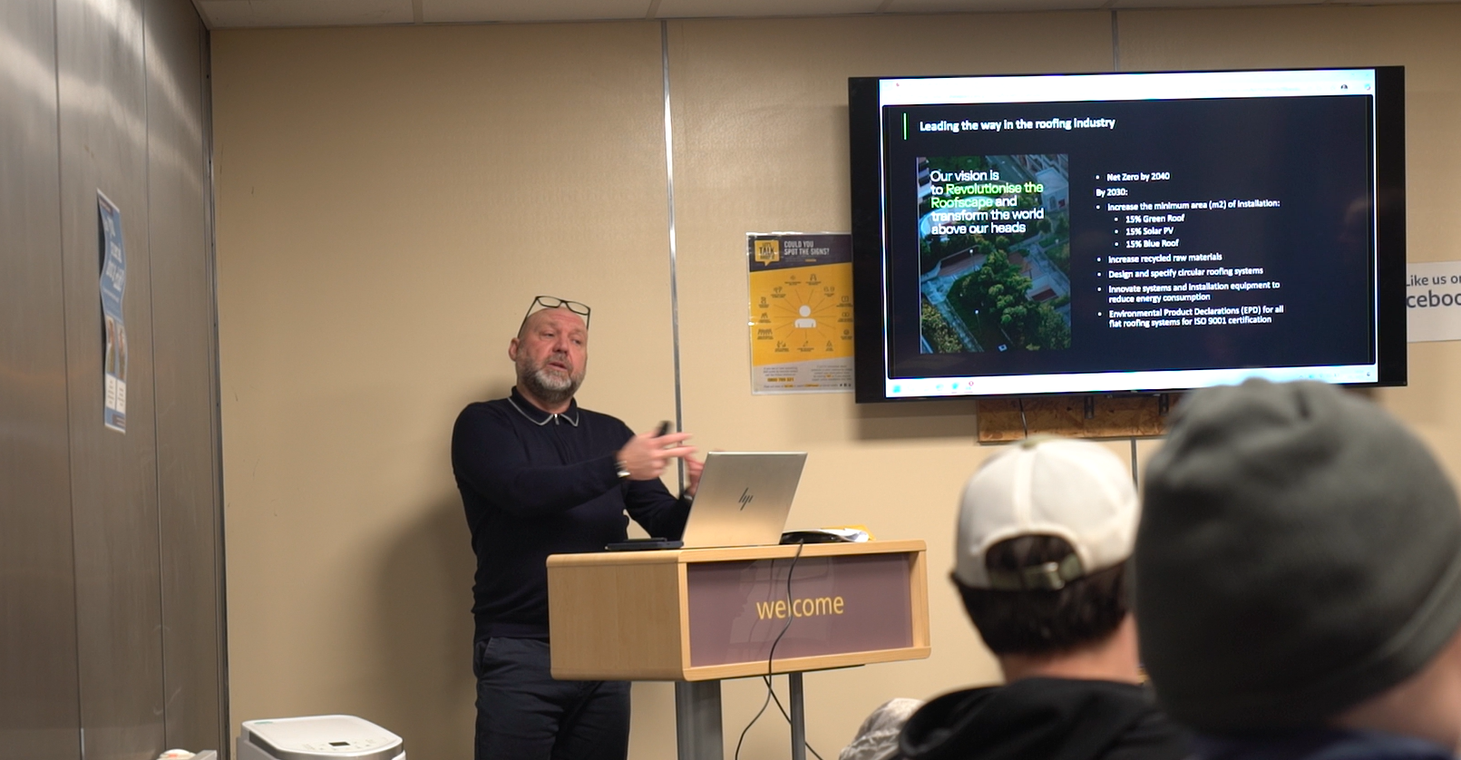 Man presenting at wooden podium with pointer, large screen showing satellite imagery, audience members visible from behind.