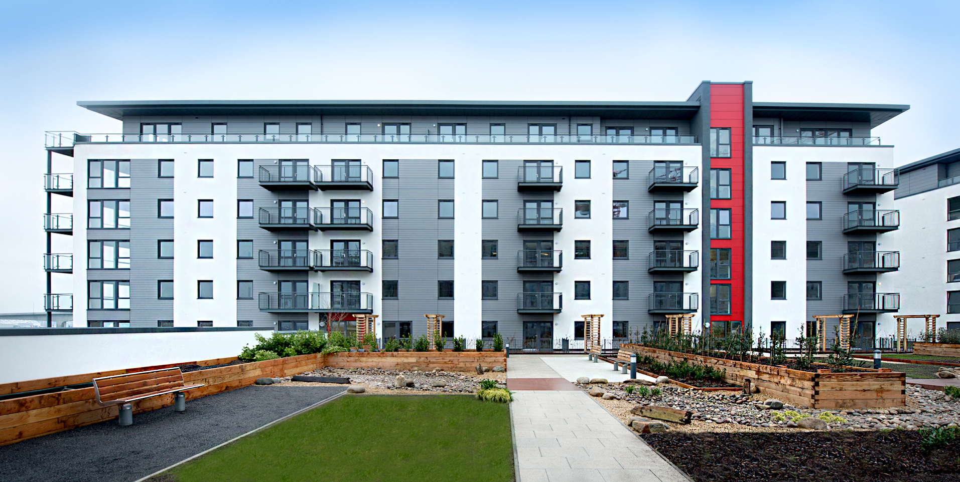 Modern apartment block with grey facade, red accent panel, and multiple balconies. Landscaped front garden with lawn and stone features.