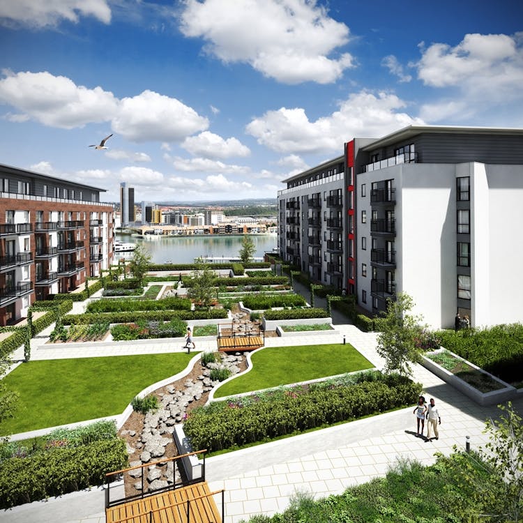 Modern residential development with grey and white apartment blocks surrounding landscaped courtyard featuring green lawns, pathways, and water view beyond under blue cloudy sky.