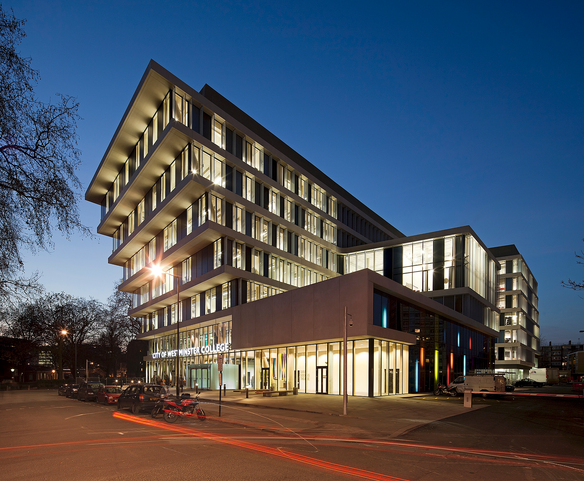 Modern multi-storey building with glass facades and warm interior lighting against deep blue evening sky, with illuminated ground floor retail space.