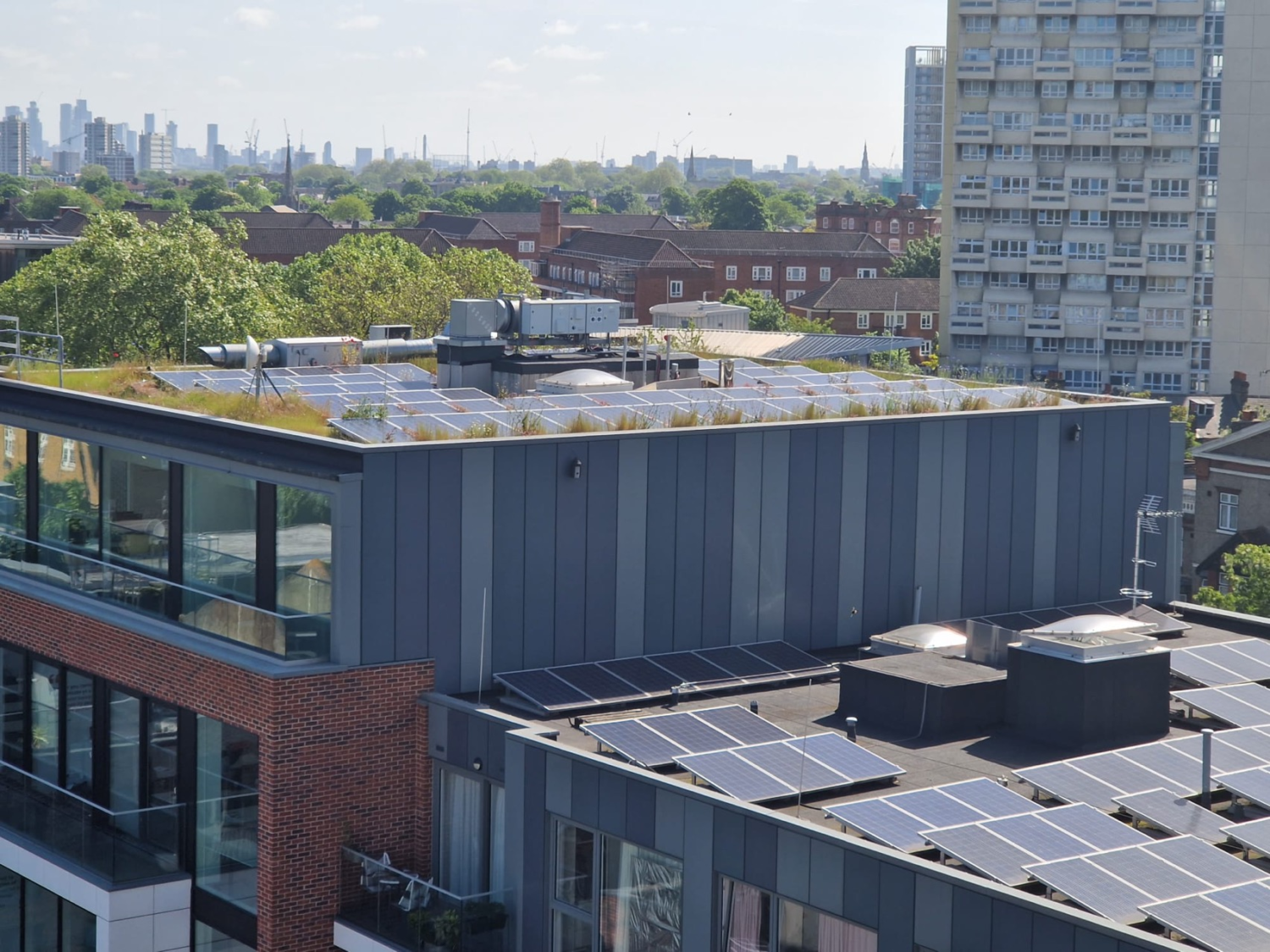 Modern multi-storey building with dark vertical cladding, solar panels on lower roof, brick base, surrounded by urban landscape with trees and high-rise blocks in distance.