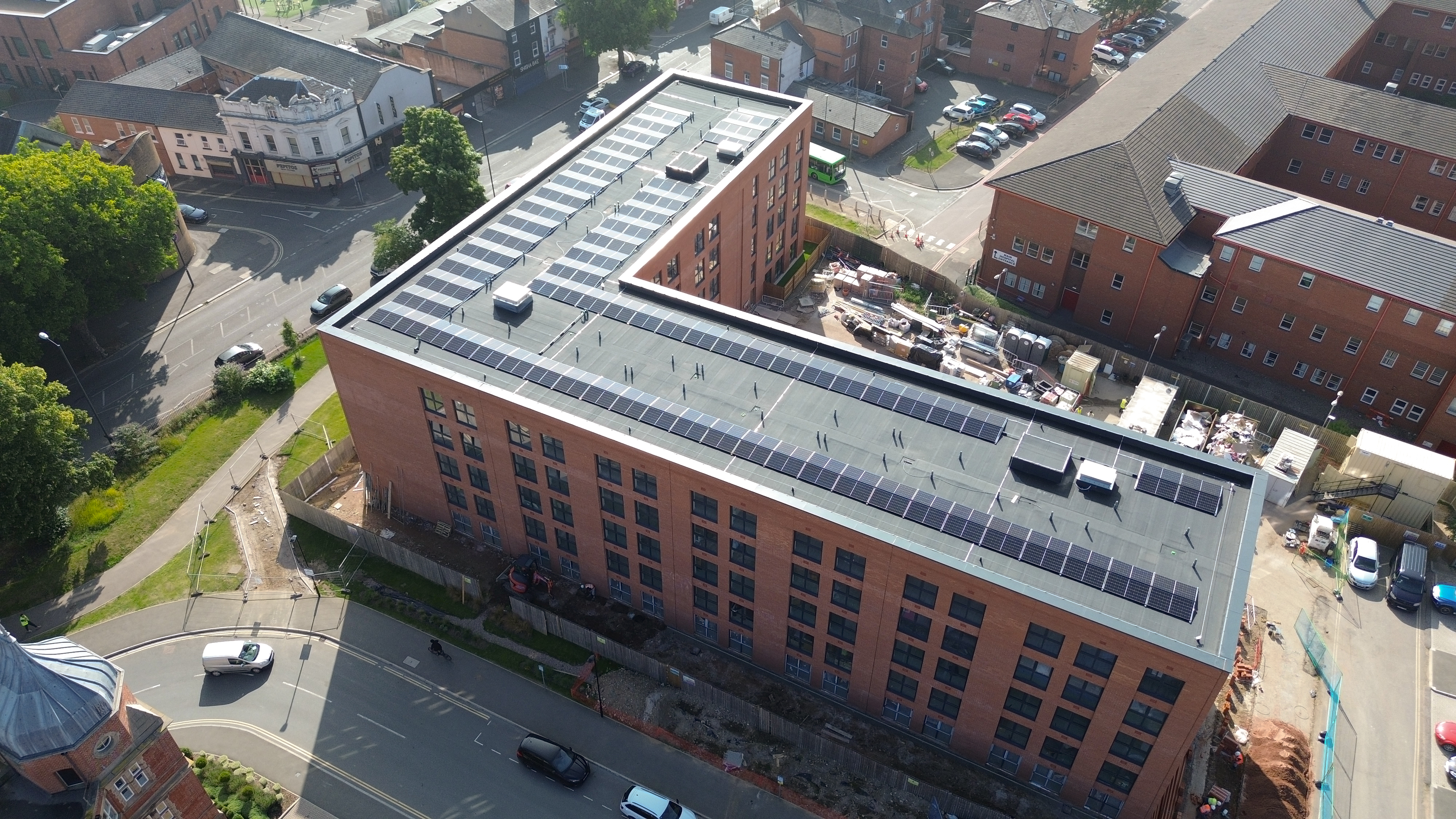 Aerial view of modern brick building with glass roof structure, surrounded by residential houses and streets with parked cars.