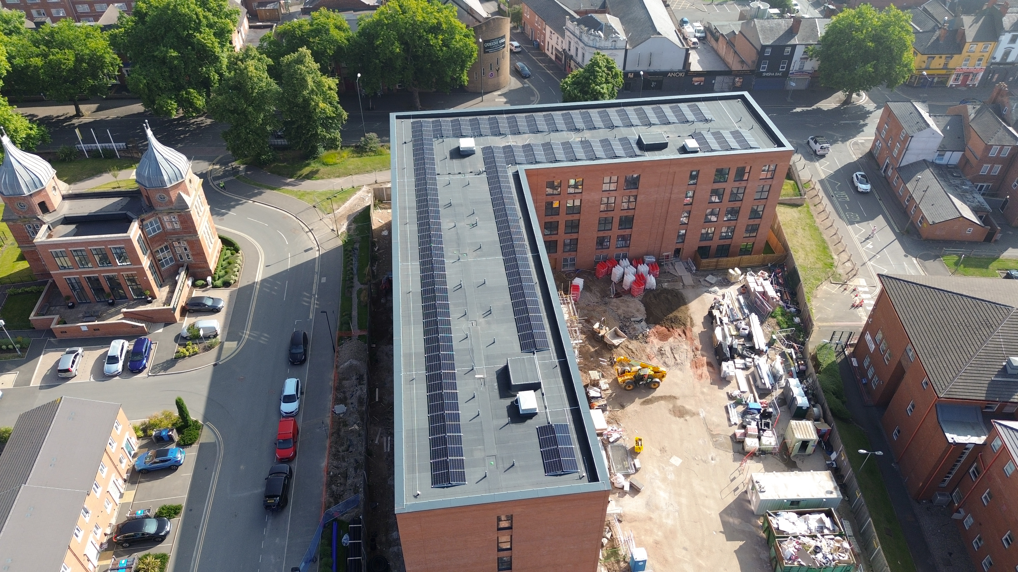 Aerial view of brick building with grey metal roof under construction, surrounded by residential houses, trees, and parked cars.