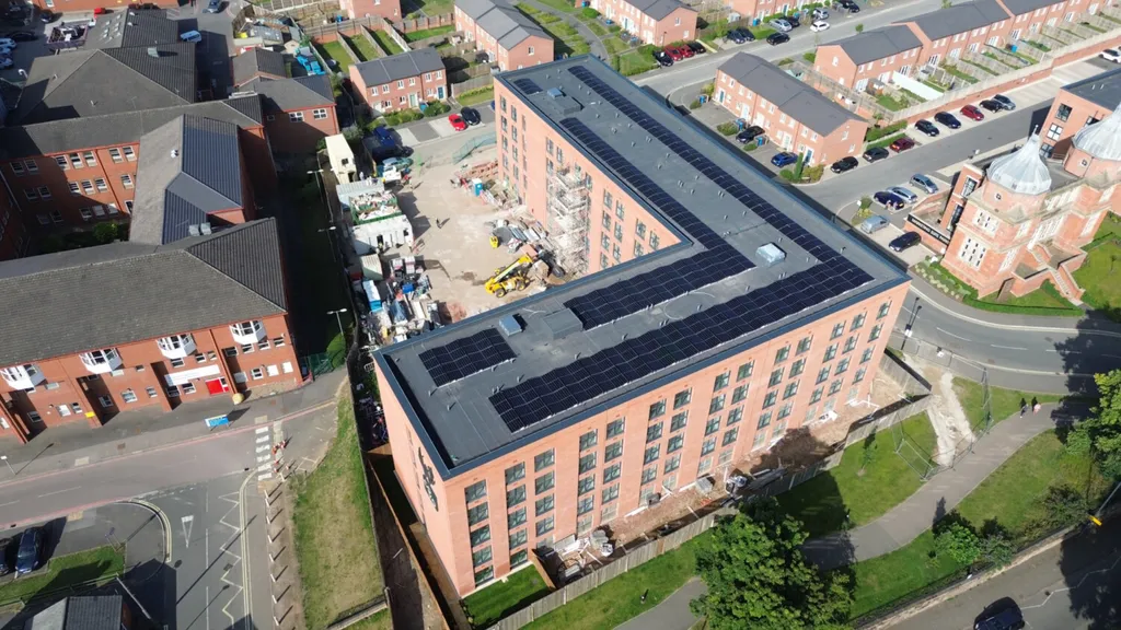 Aerial view of modern brick apartment building with dark roof surrounded by residential housing estate and roads.