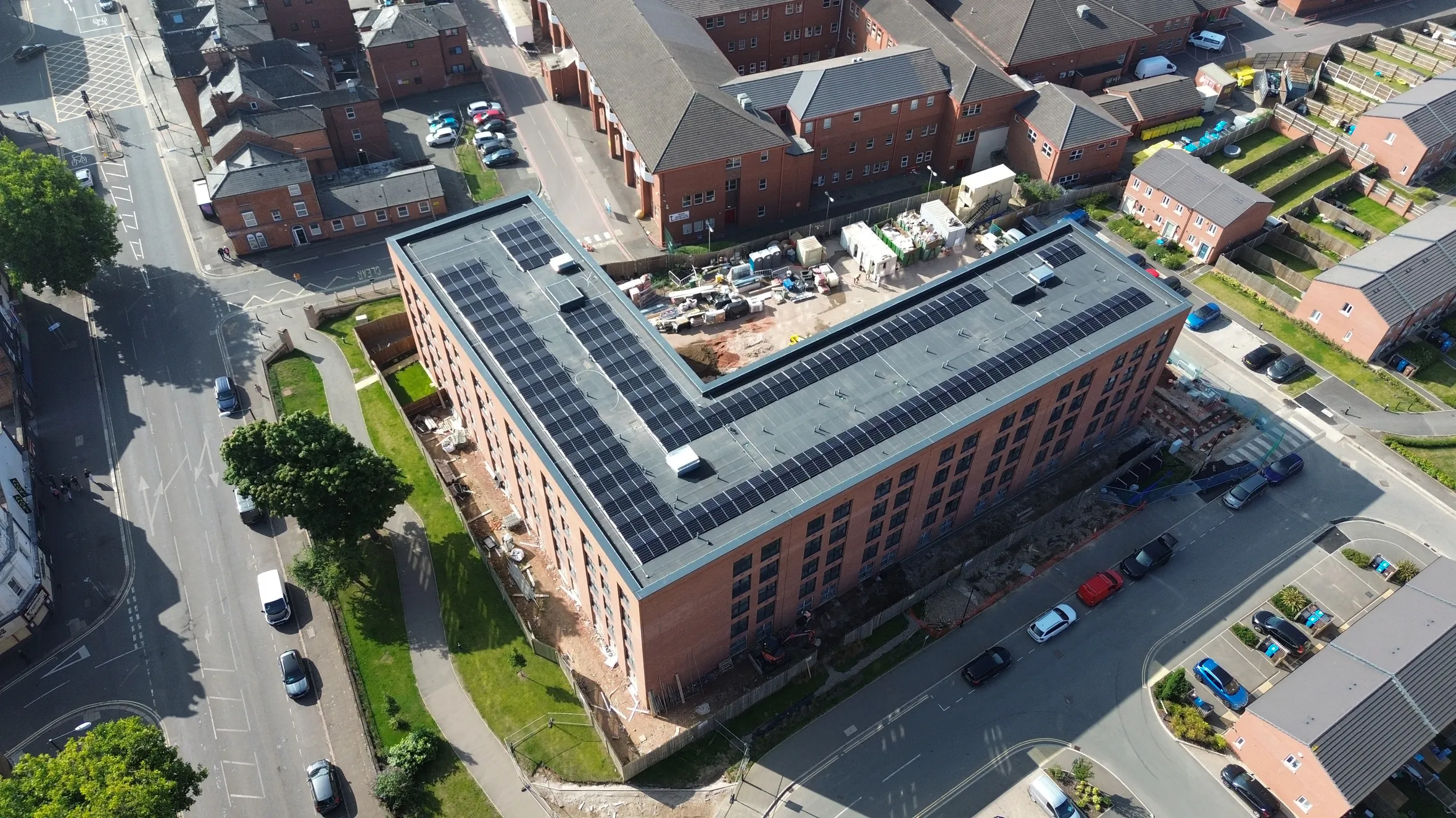Aerial view of large brick building with solar panels on flat roof, surrounded by residential houses, car parks, and tree-lined streets.