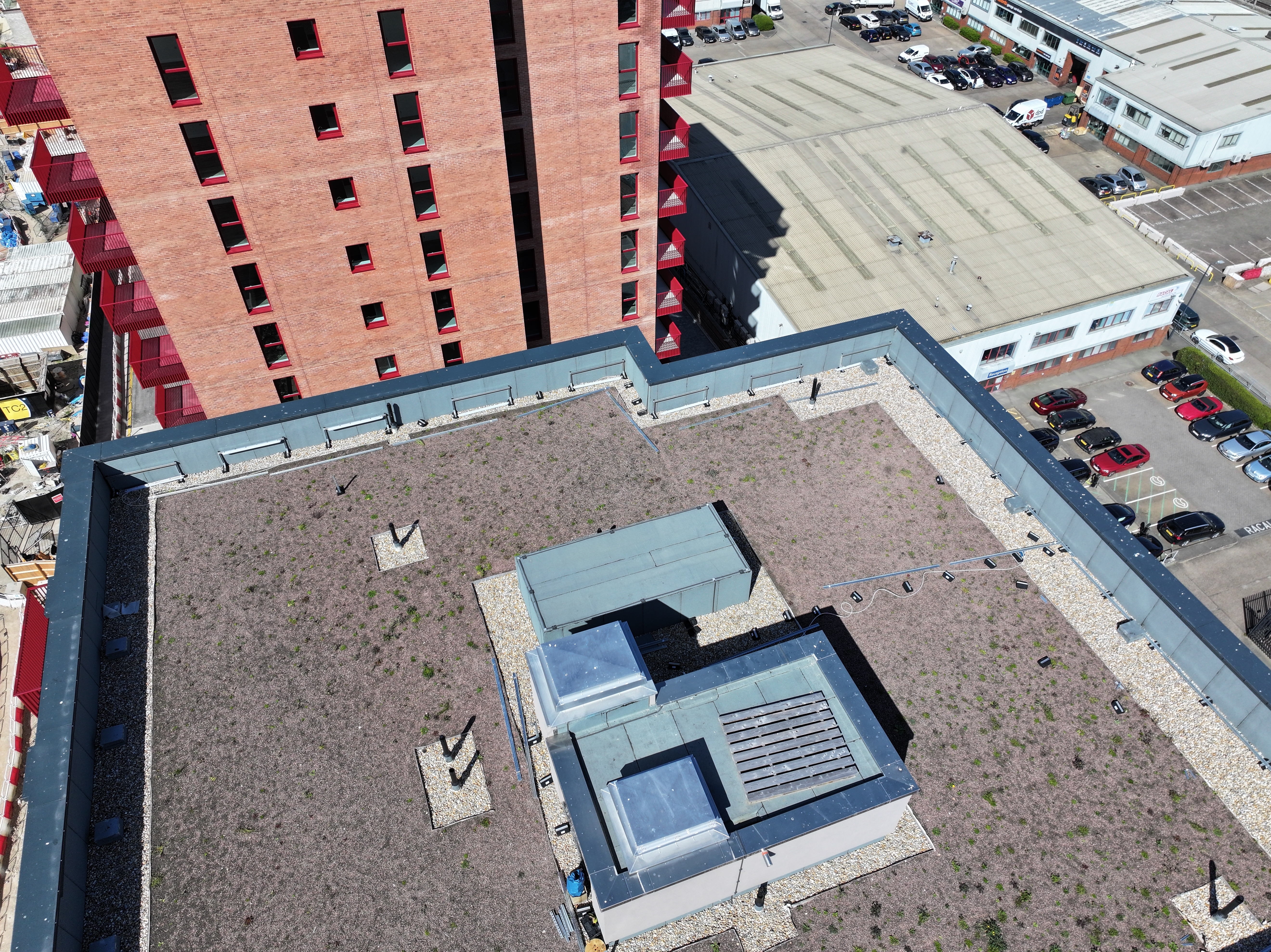 Aerial view of modern building with grey roof, central courtyard, adjacent red brick residential tower and car parks.