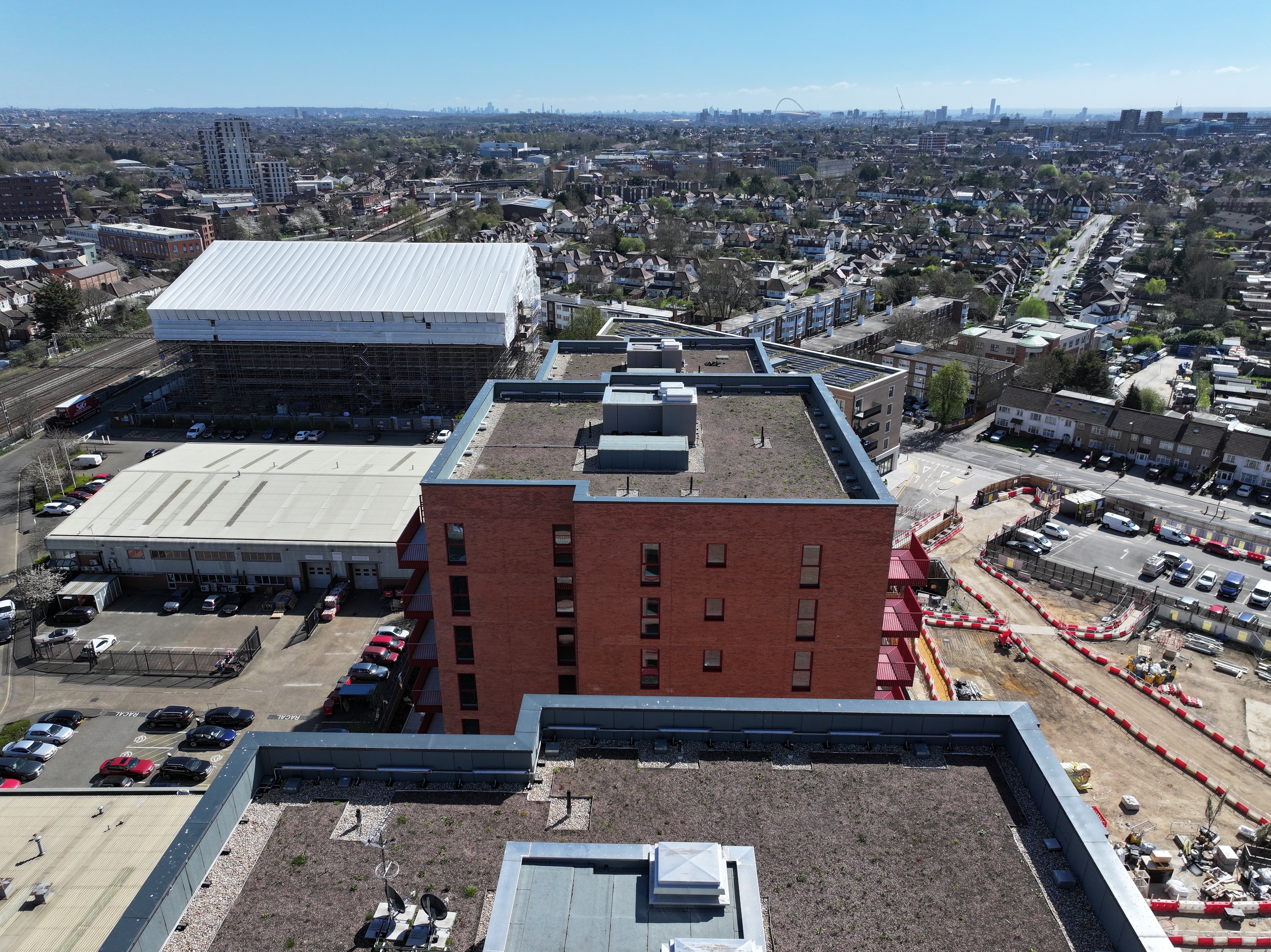 Aerial view of urban area with large red brick building, white rooftops, car parks, and residential neighbourhoods extending to horizon.