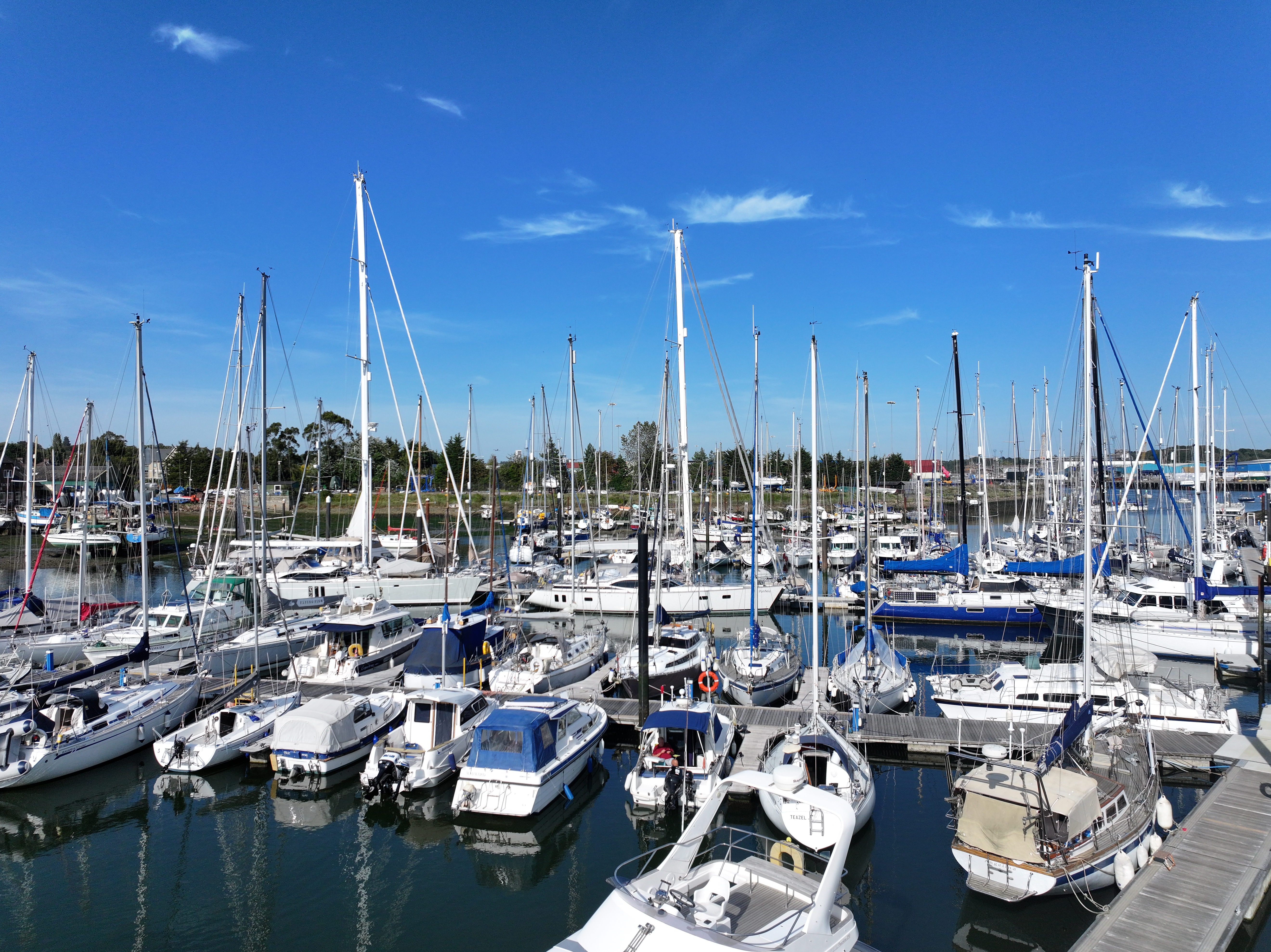 Marina filled with white sailboats and yachts with tall masts under bright blue sky with scattered white clouds.