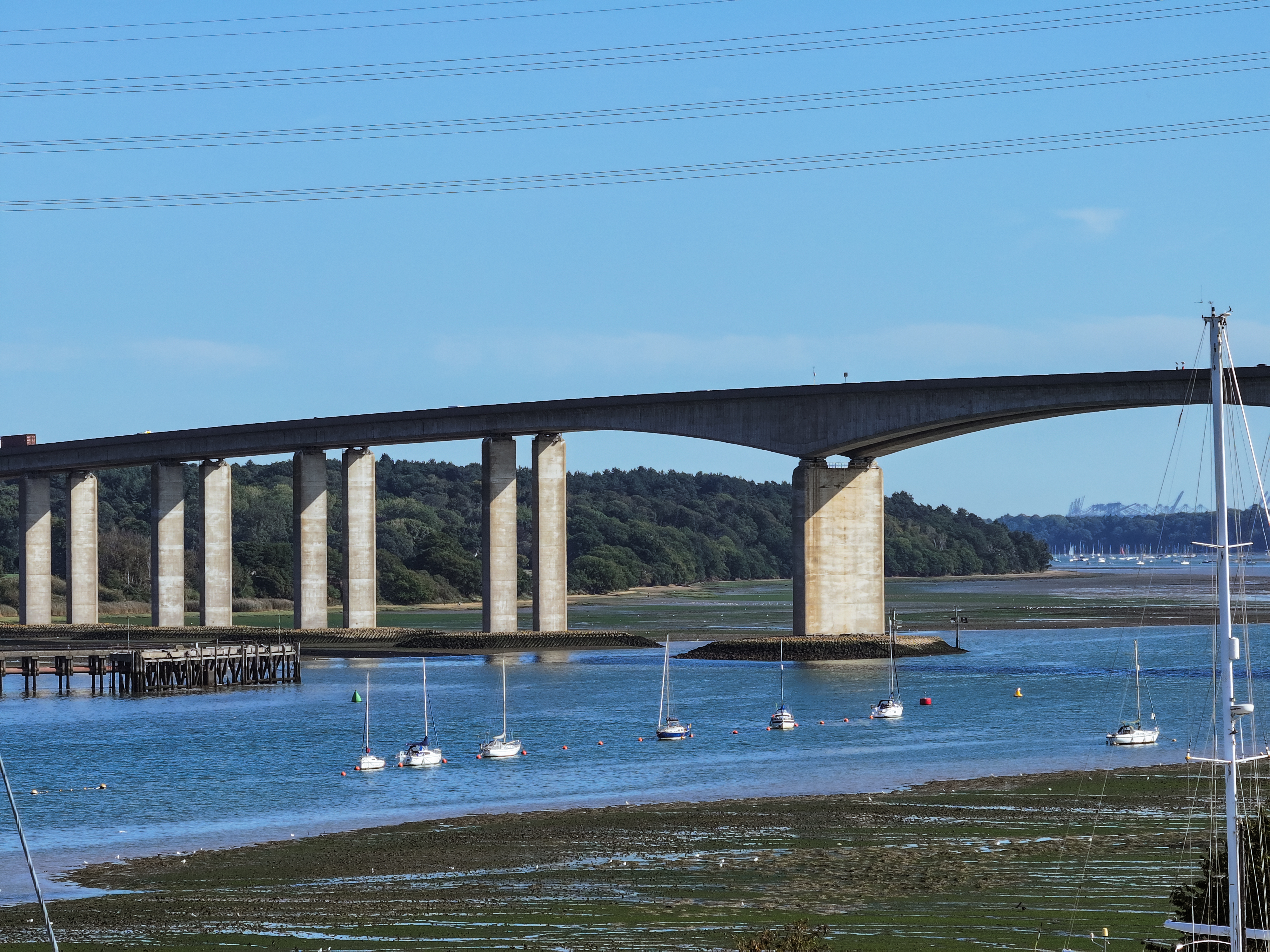 Large concrete bridge with multiple arches spanning blue water, with white sailing boats moored below and green hills in background.