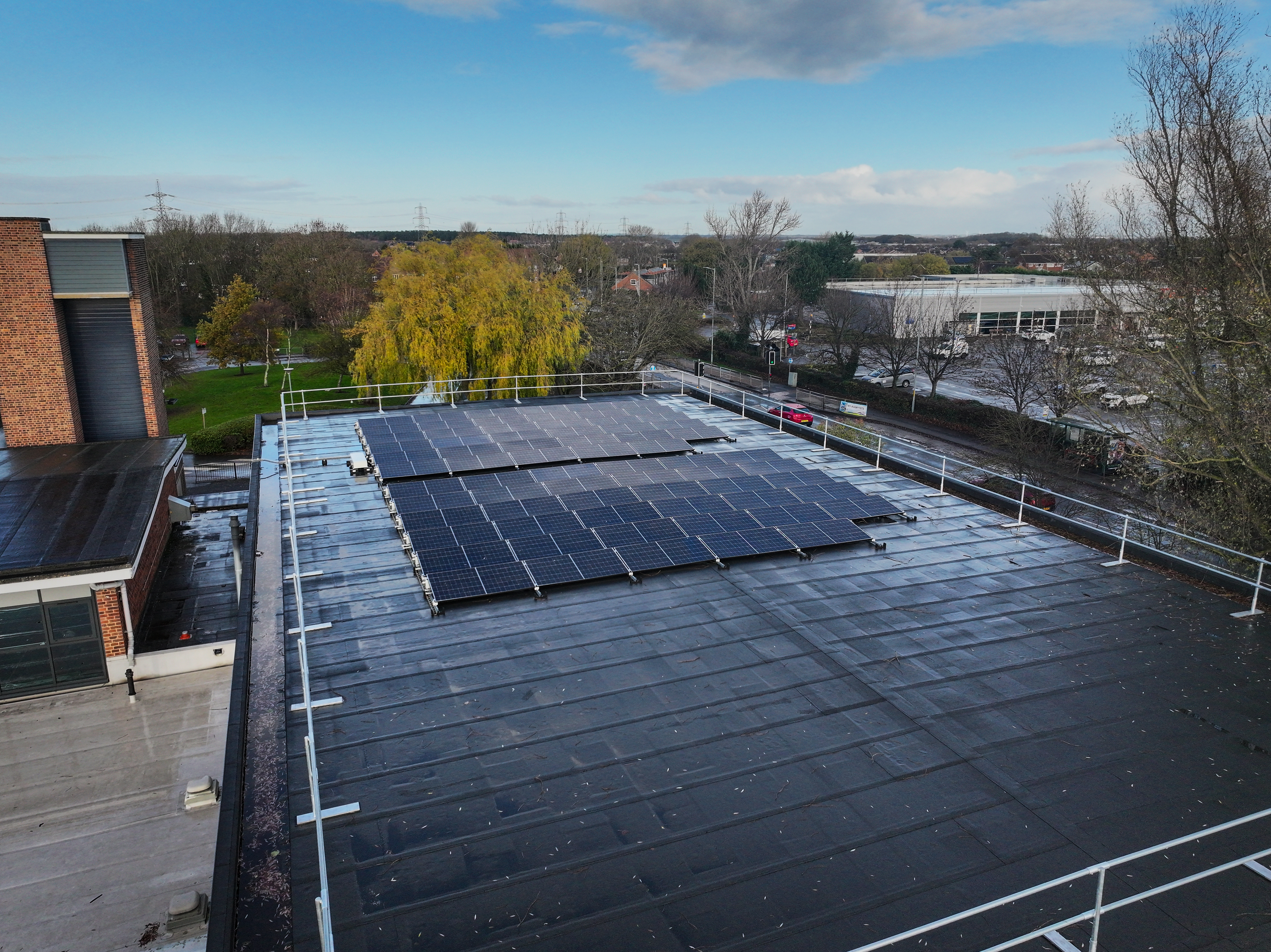 Aerial view of dark solar panels installed on flat rooftop with white safety railings, surrounded by trees and buildings under blue sky