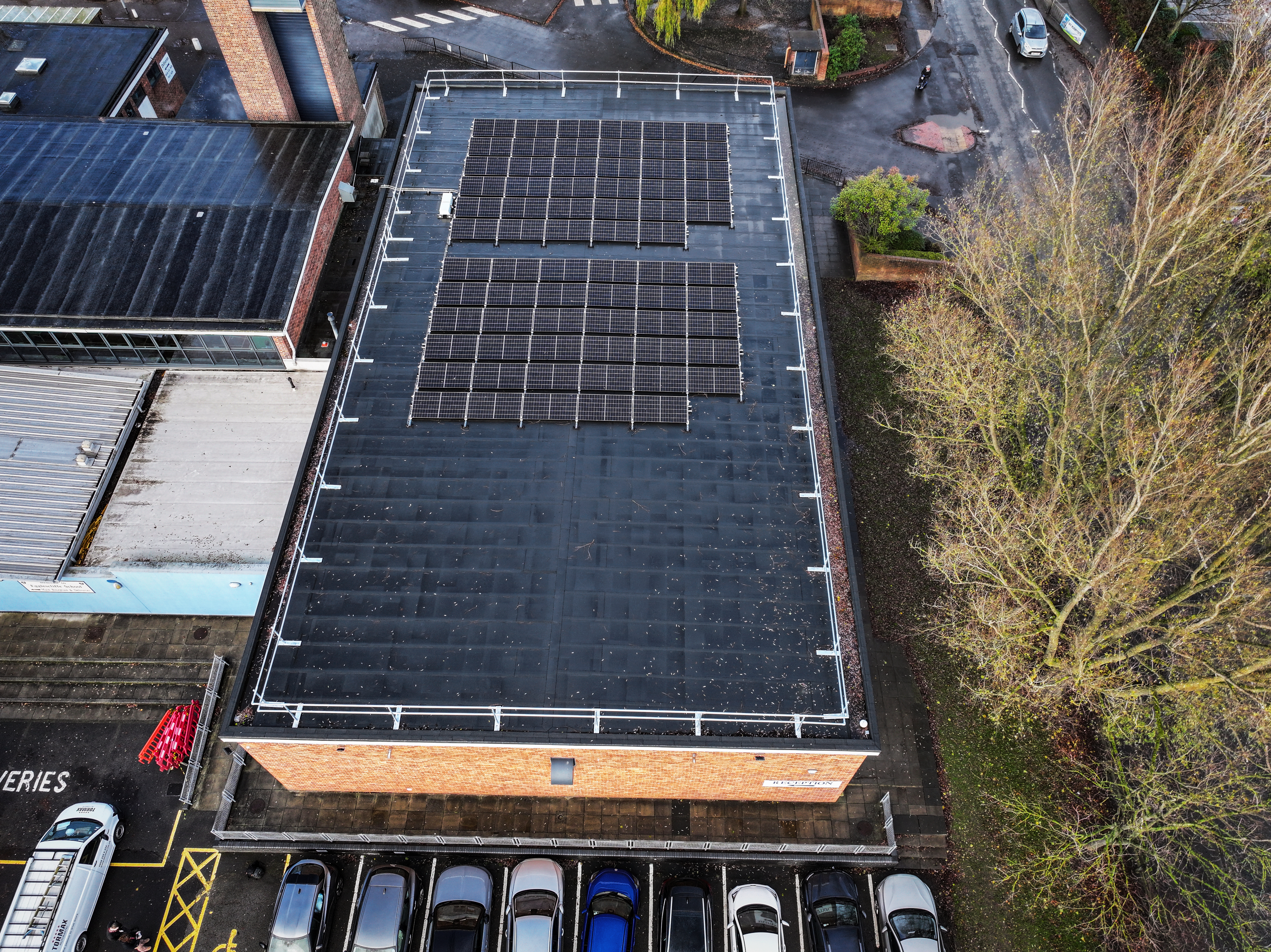 Aerial view of flat-roofed building with solar panels arranged in rows on dark roof surface, cars parked below, bare trees visible