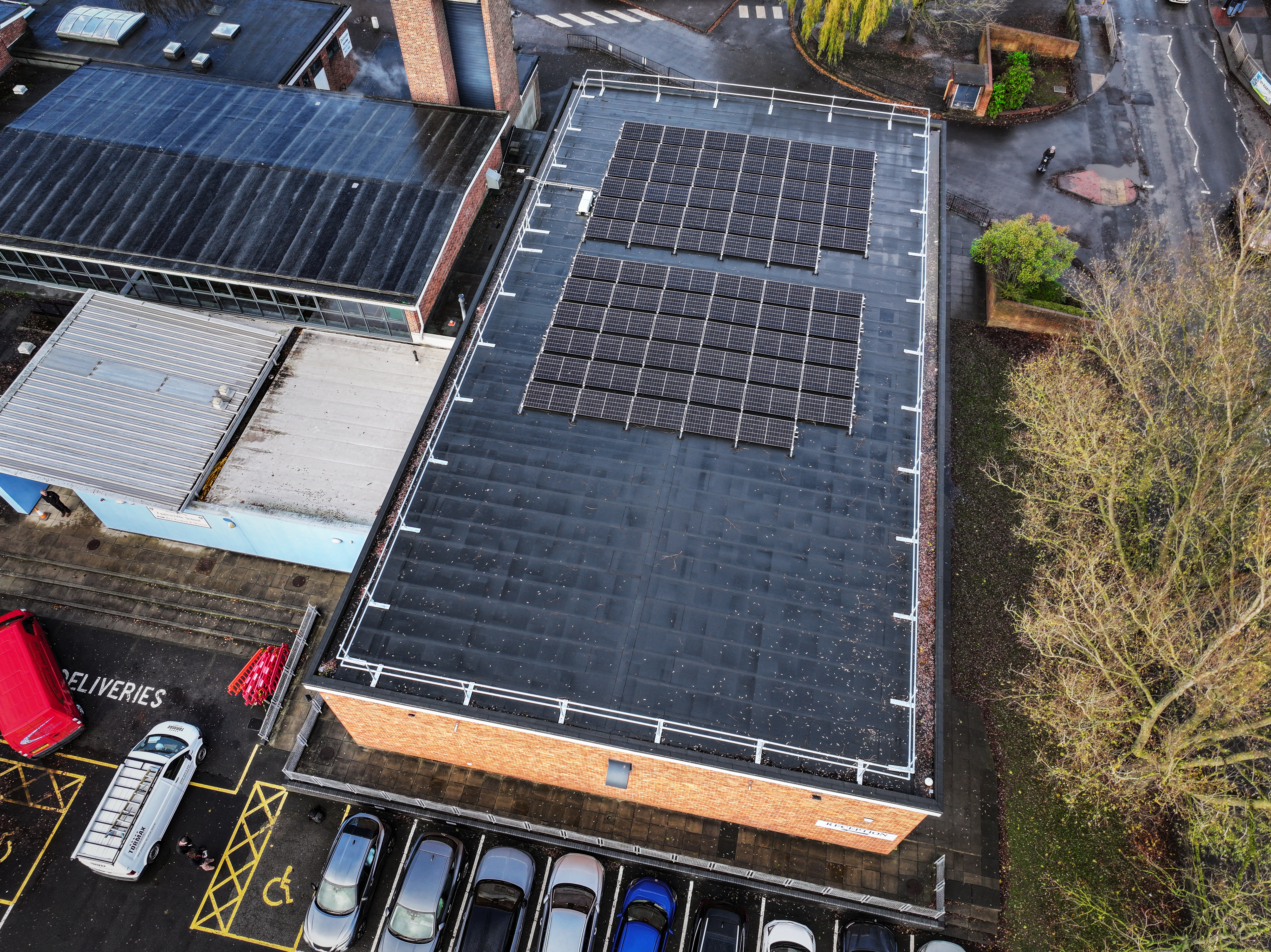 Aerial view of industrial building with dark solar panels covering large flat roof, cars parked below, surrounding buildings visible