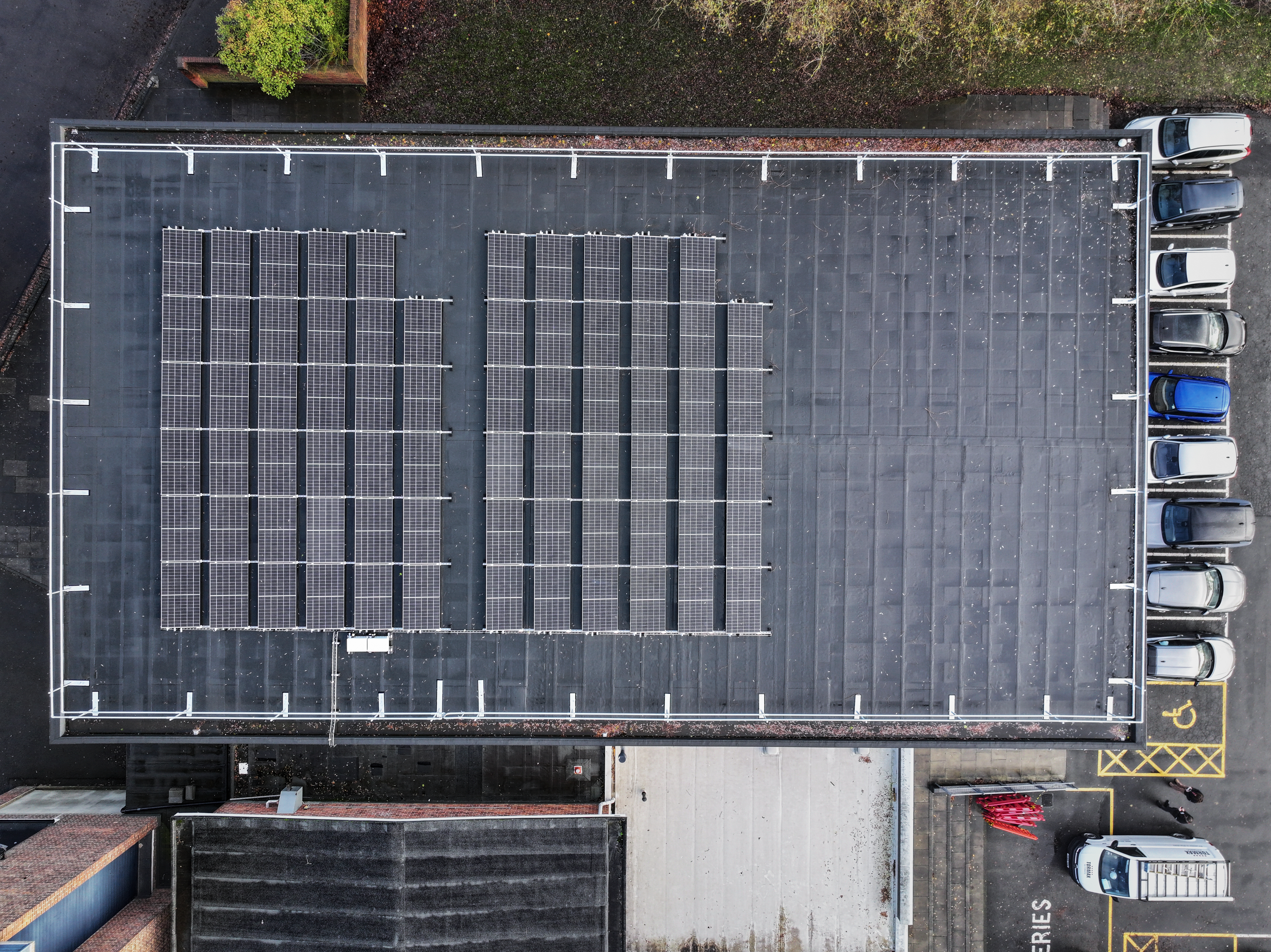 Aerial view of rectangular grey rooftop with dark solar panel arrays arranged in grid patterns, surrounded by white railings and parked cars