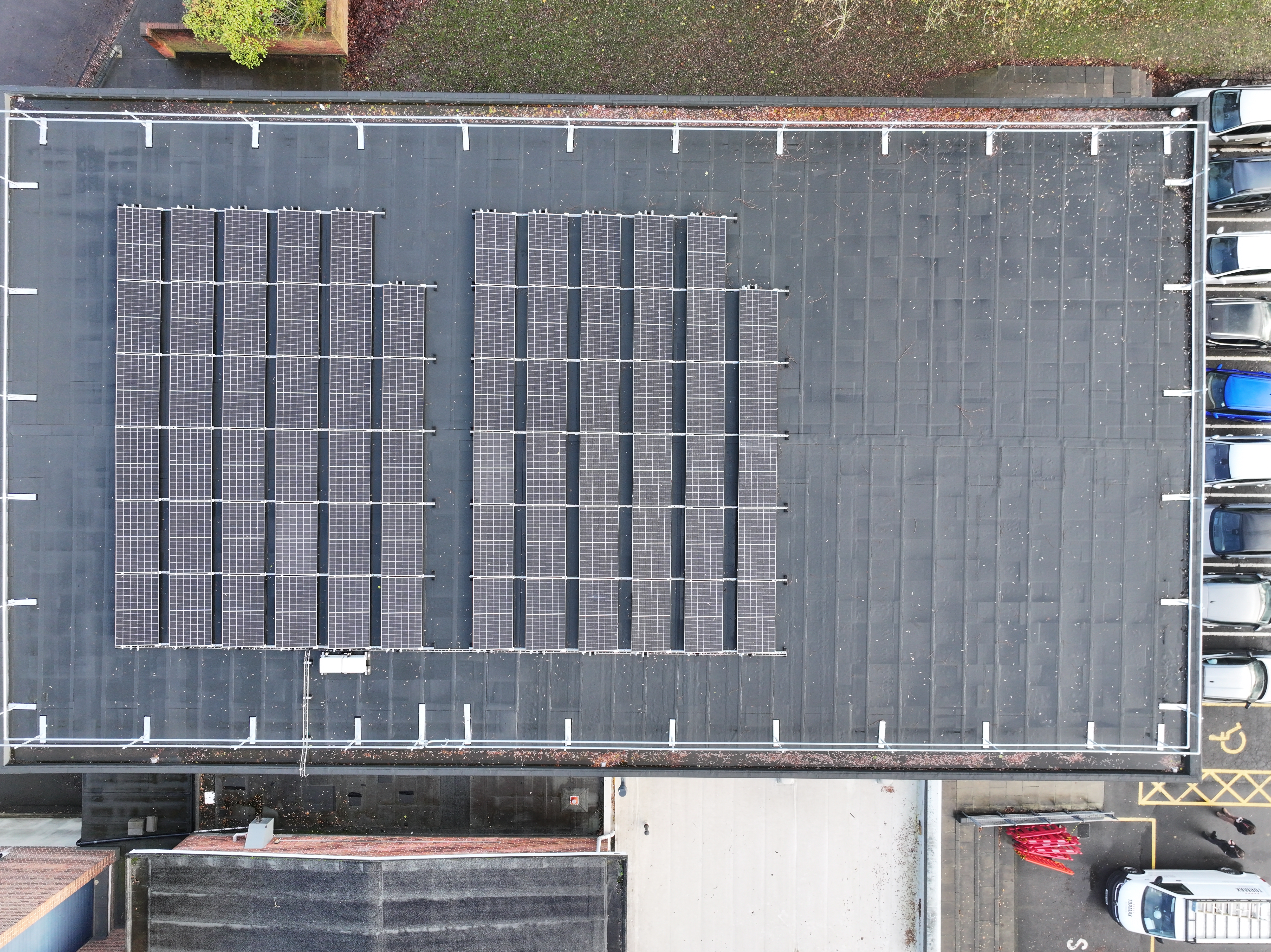 Aerial view of grey warehouse roof with dark rectangular solar panels arranged in two groups, surrounded by car park and buildings