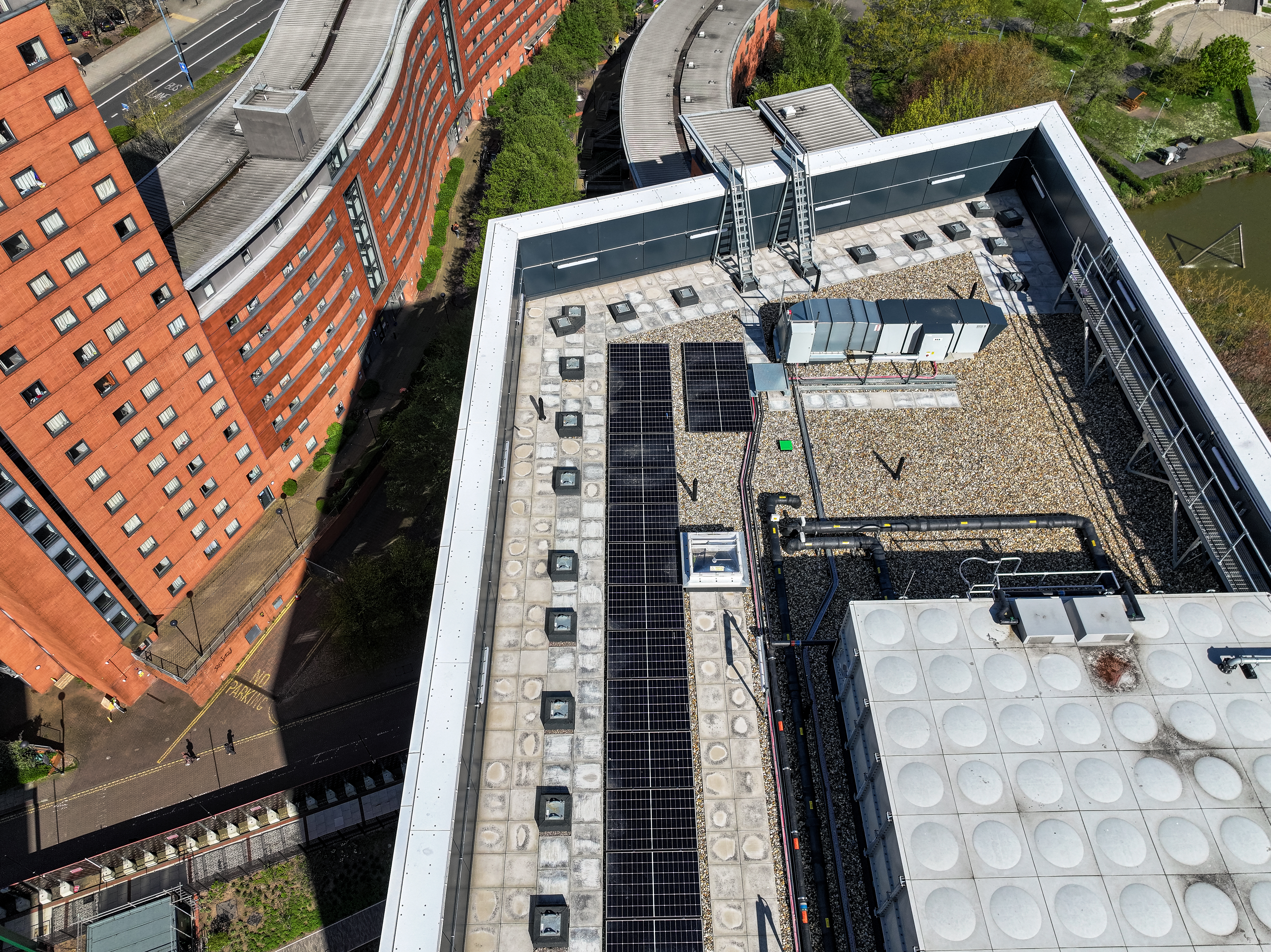 Aerial view of a flat rooftop with solar panels, HVAC units, and gravel surface, next to a curved red brick building and road.