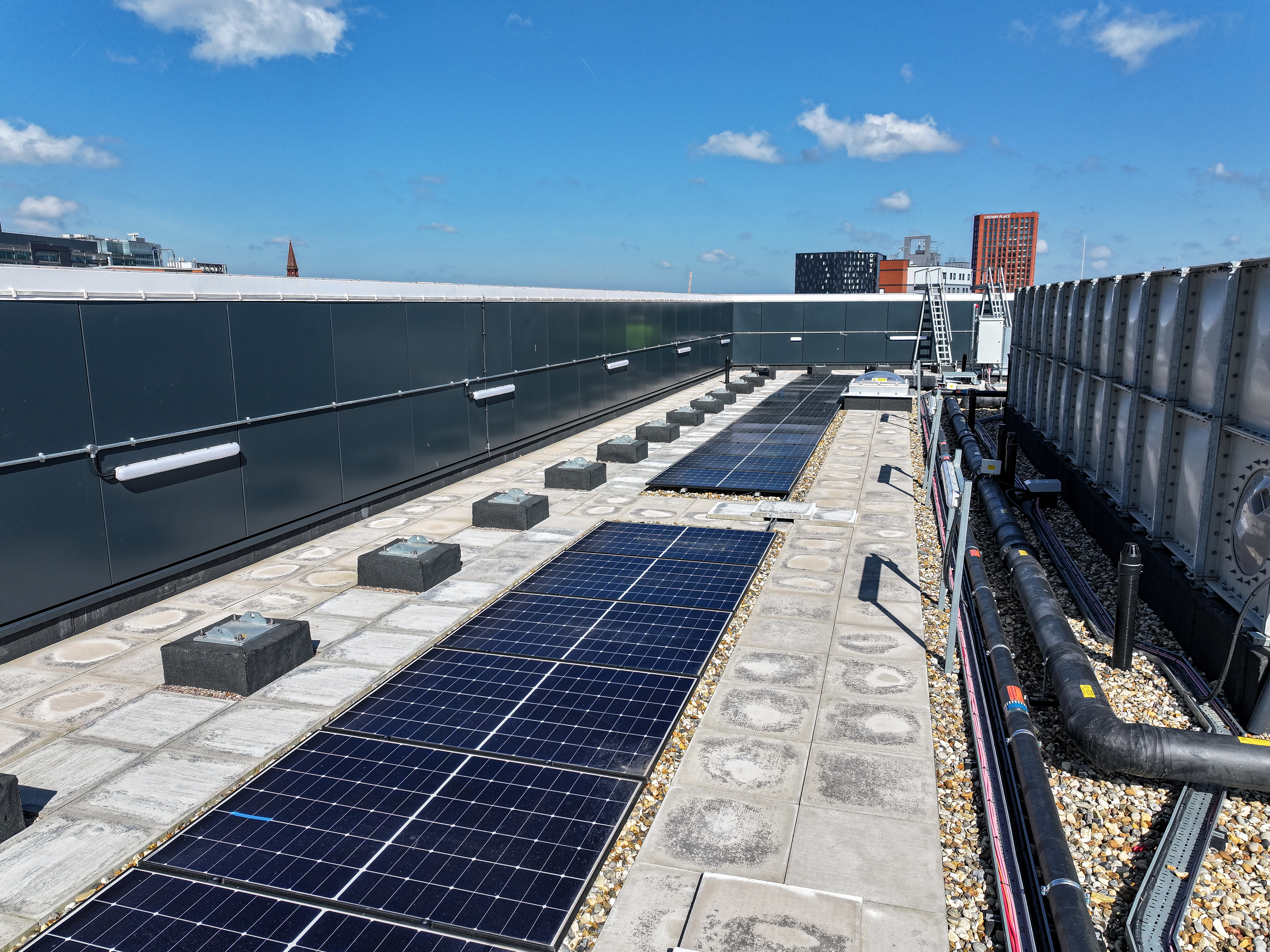 Solar panels installed on a flat urban rooftop with gravel surface, pipework, and ventilation units under a blue sky.