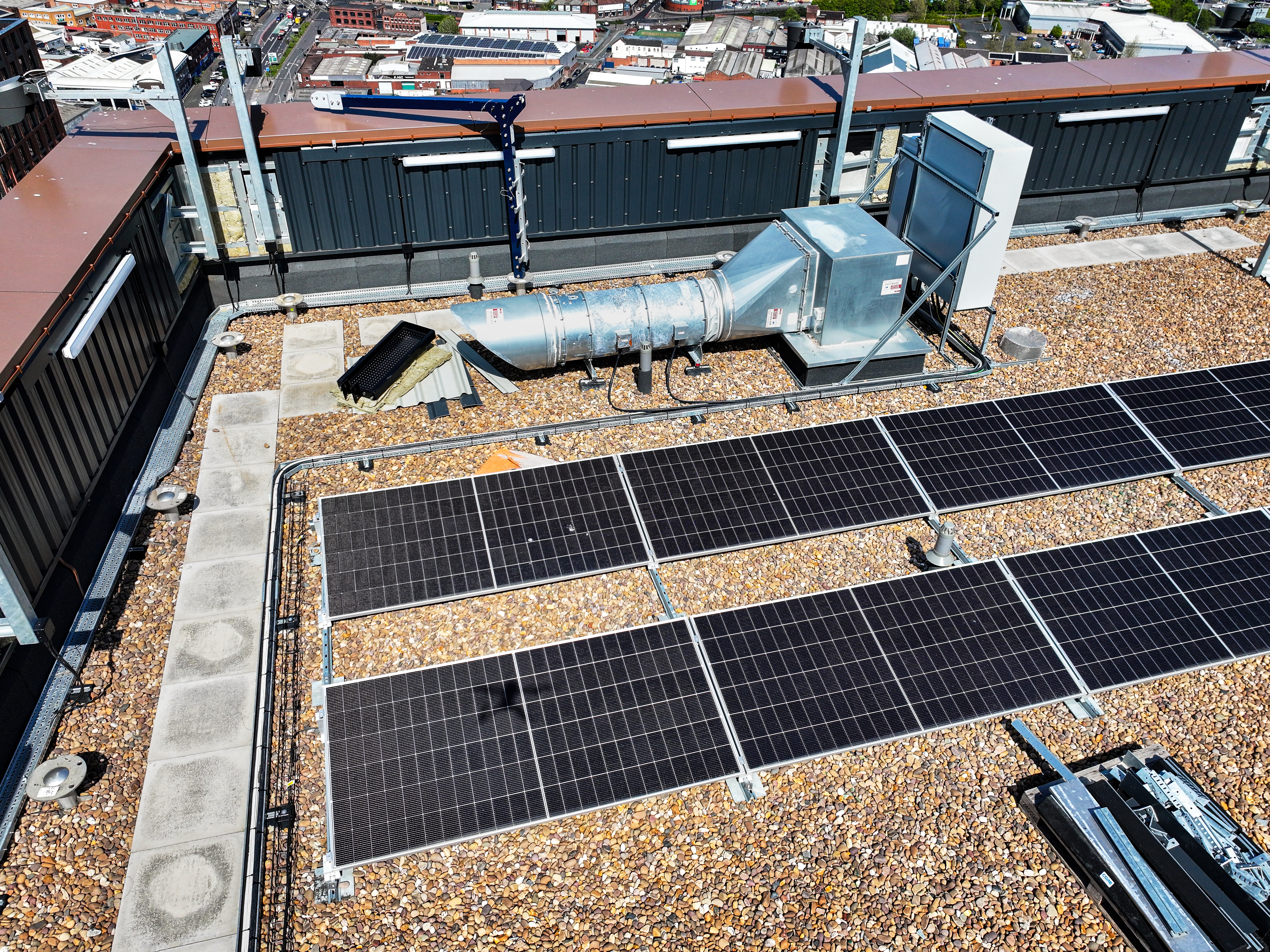 Aerial view of a flat rooftop with solar panels on gravel, a metal ventilation unit, and dark-clad buildings in the background.