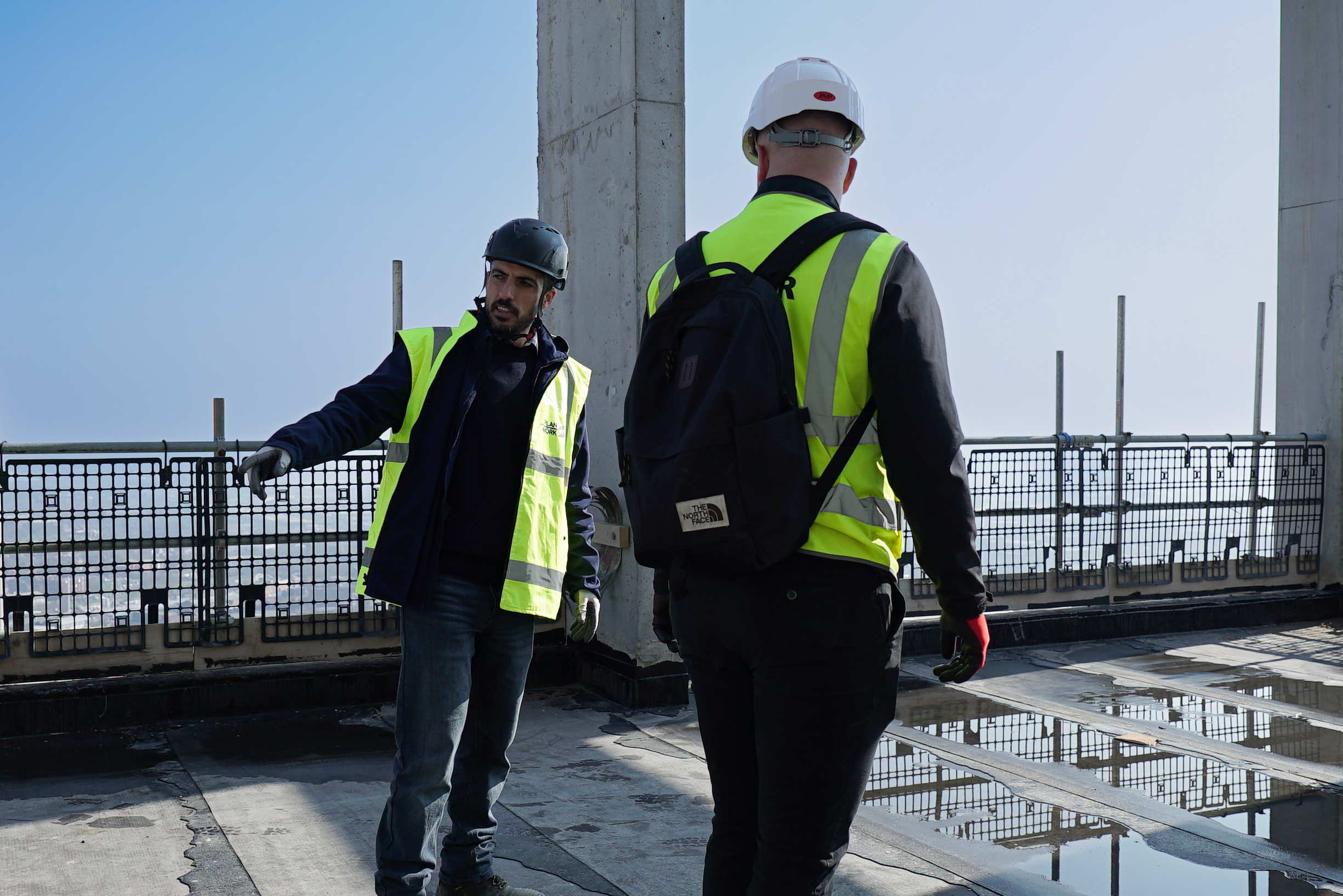 Two construction workers in hi-vis yellow vests and hard hats walking on concrete structure with metal railings and pillars.