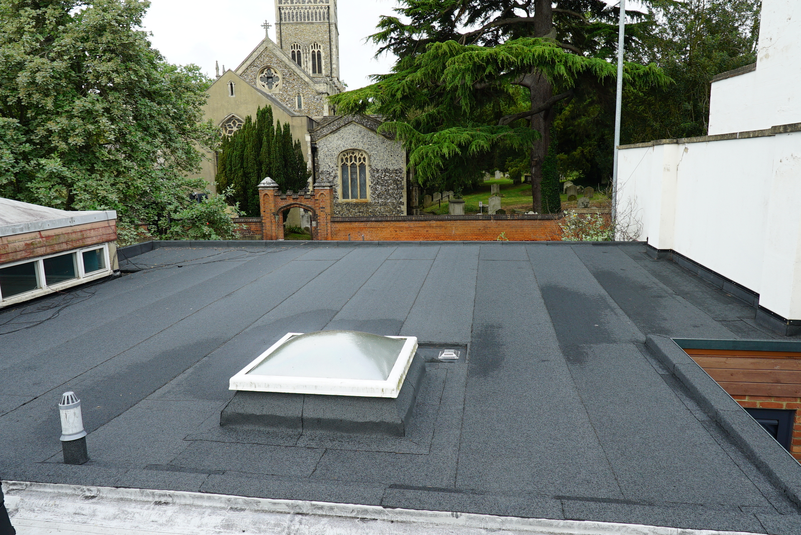Flat roof with dark membrane covering, white rectangular skylight, and Gothic church with tower visible beyond trees in background.