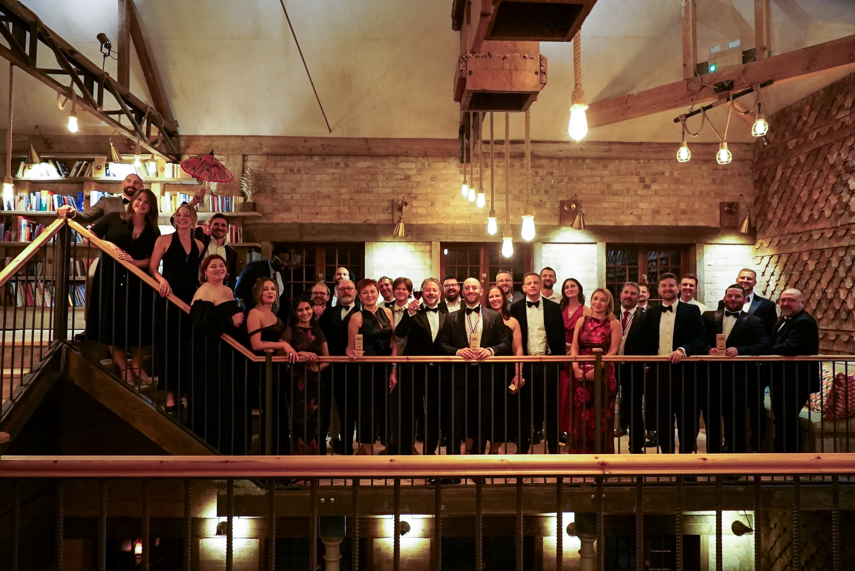 Large group in formal attire standing on upper level balcony with black railings in brick-walled venue with exposed beams and lighting.