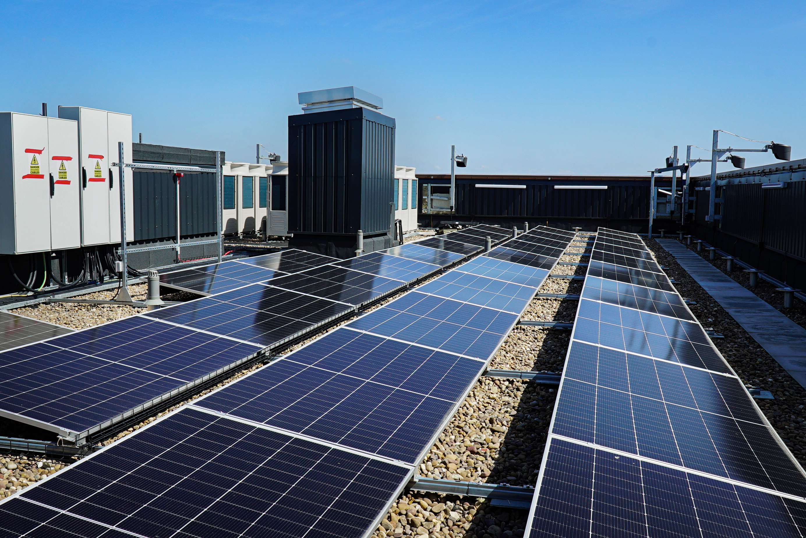 Rows of solar panels on a flat rooftop with gravel, alongside HVAC units and electrical cabinets under a clear blue sky.