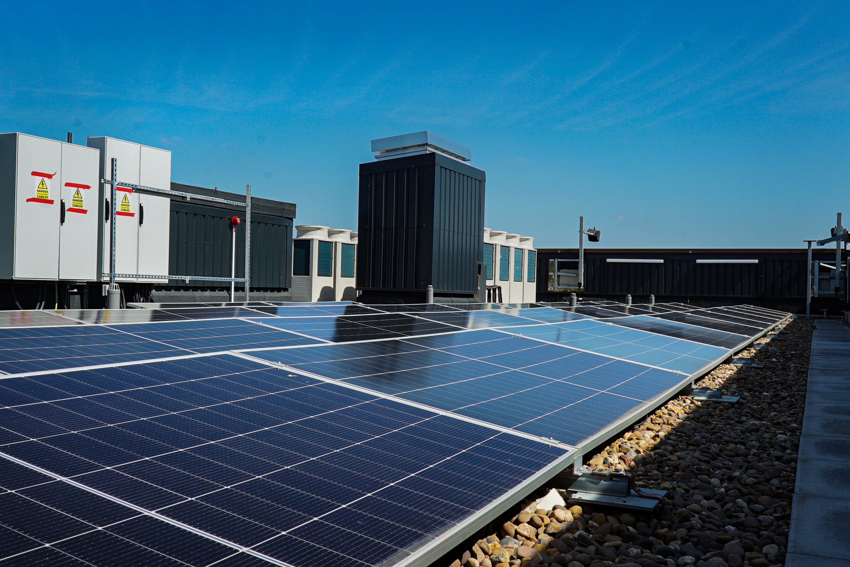 Solar panels on a flat rooftop surrounded by gravel, with HVAC units and ventilation equipment in the background under a clear blue sky.