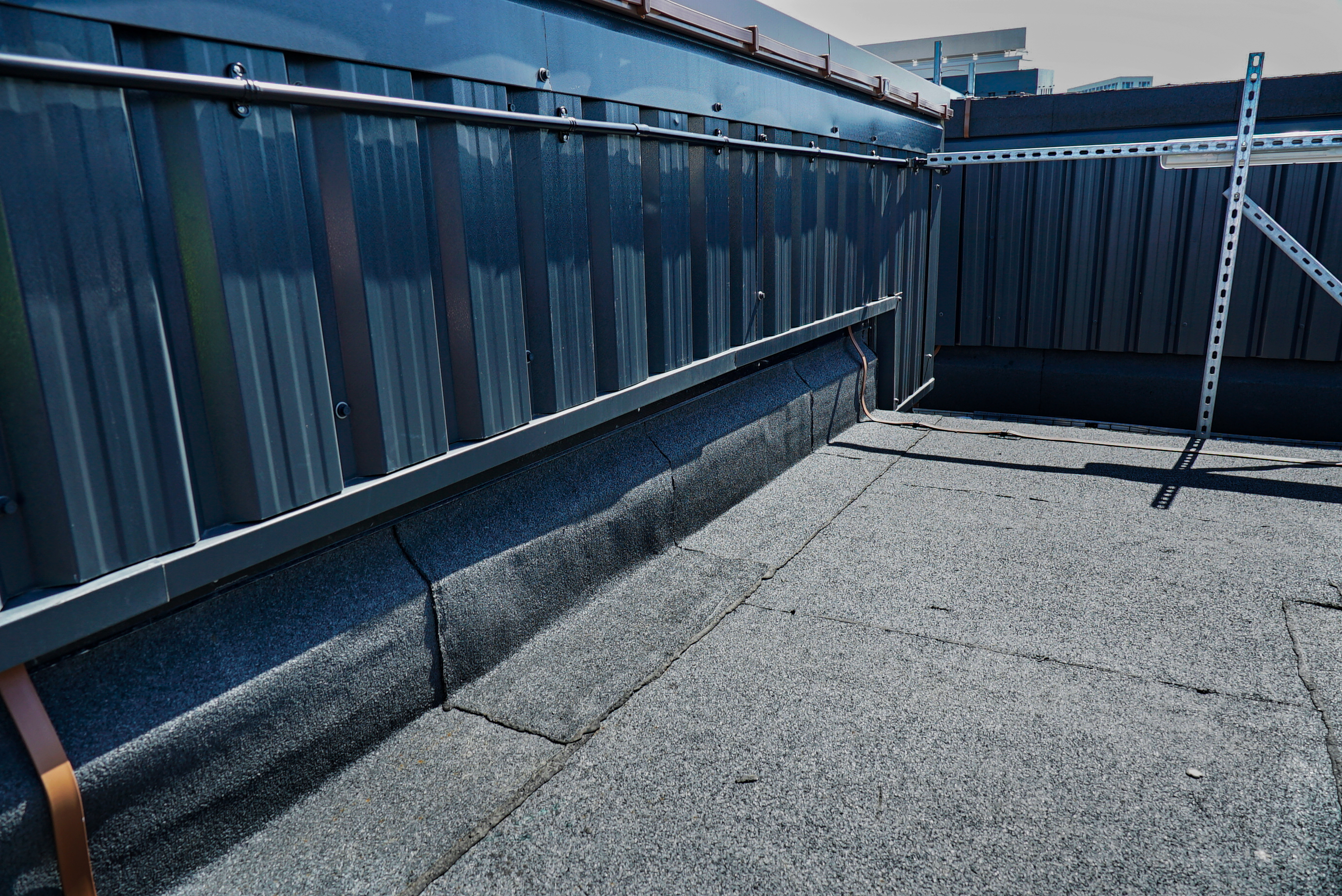 Flat rooftop with grey felt surface, surrounded by dark corrugated metal hoarding panels and a metal handrail along one side.