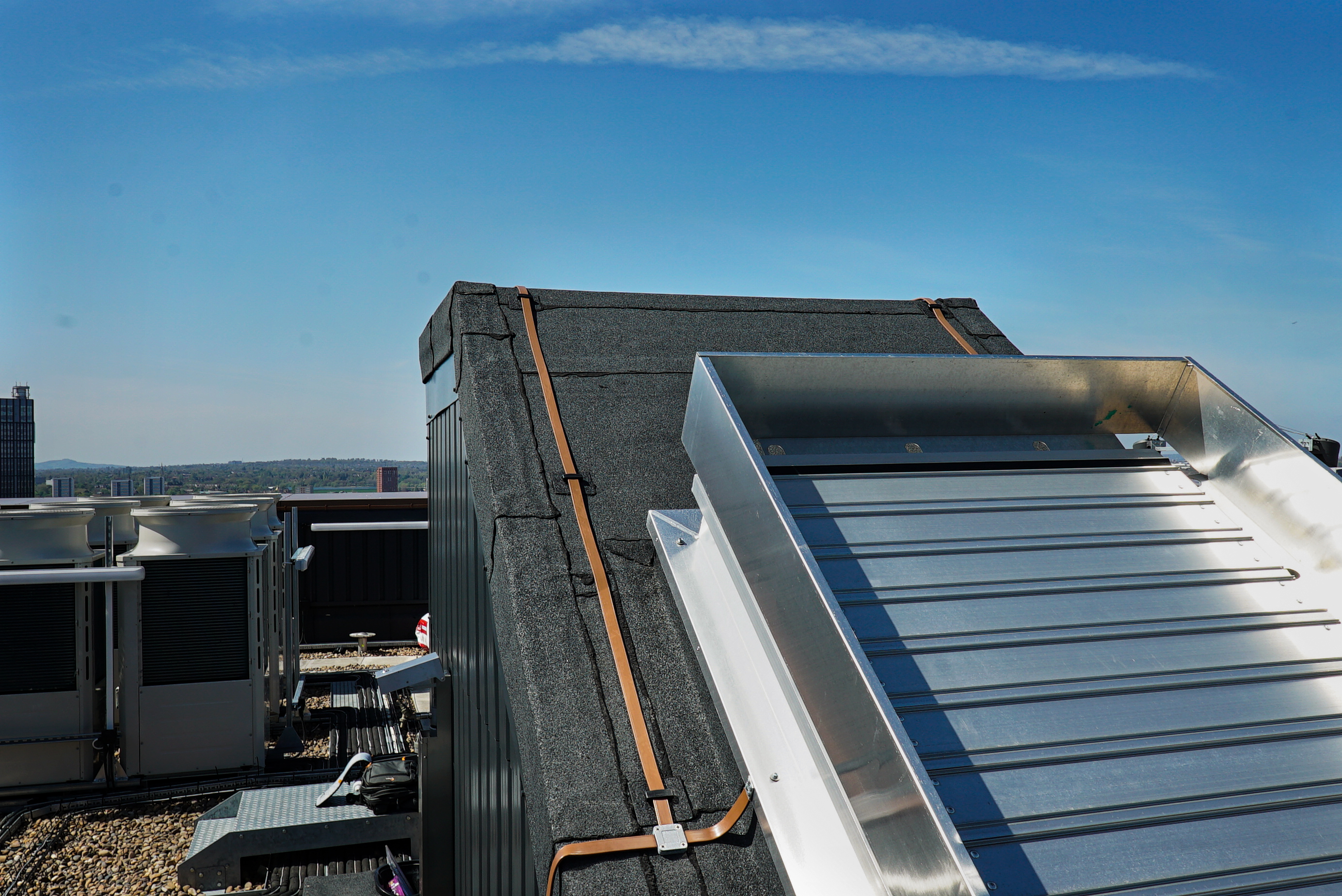 Rooftop with metal cladding, skylights, and HVAC units under a clear blue sky, with a city skyline visible in the distance.