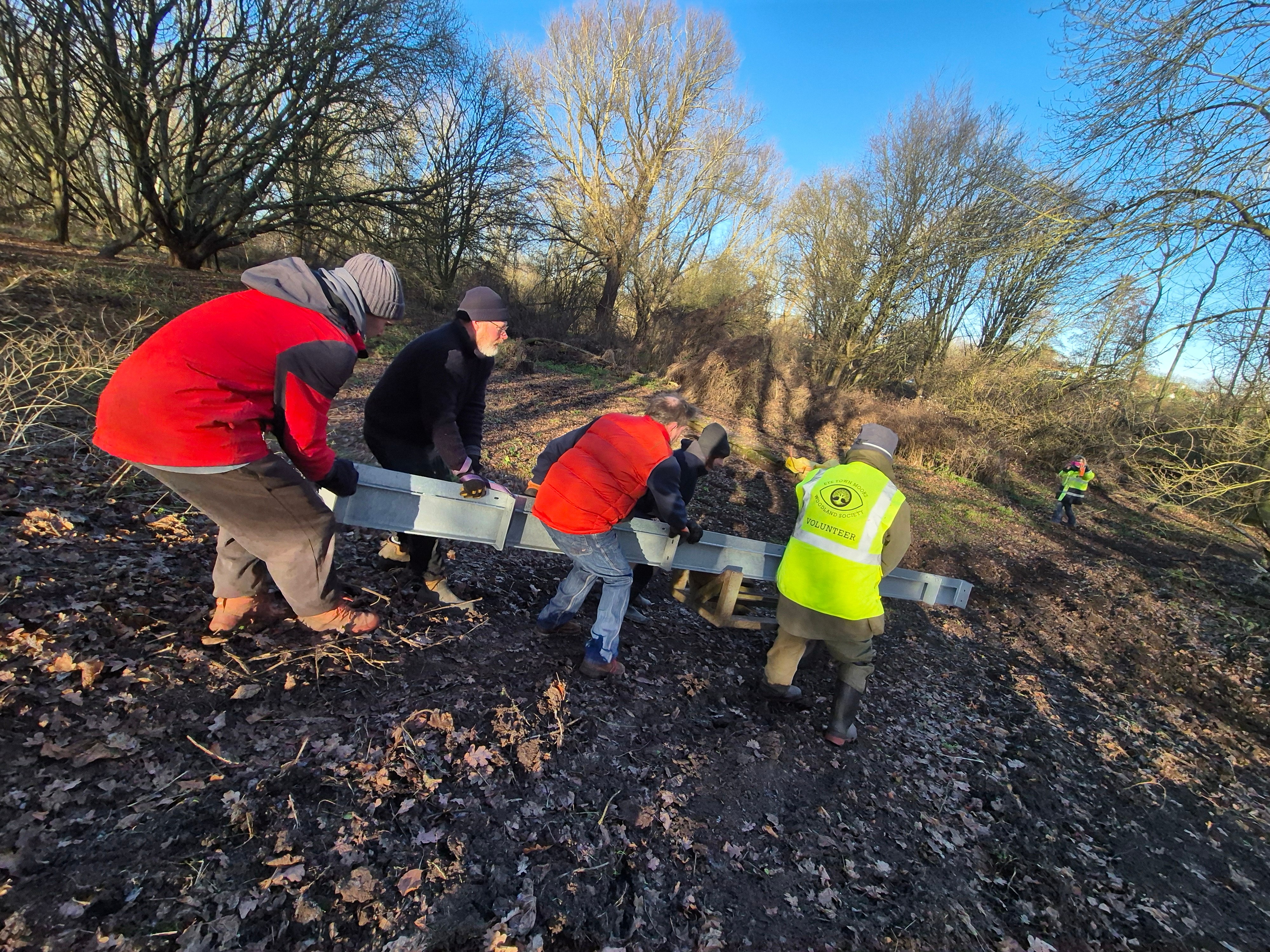Four people in red and high-vis jackets kneeling on muddy ground using tools amongst bare trees under blue sky.
