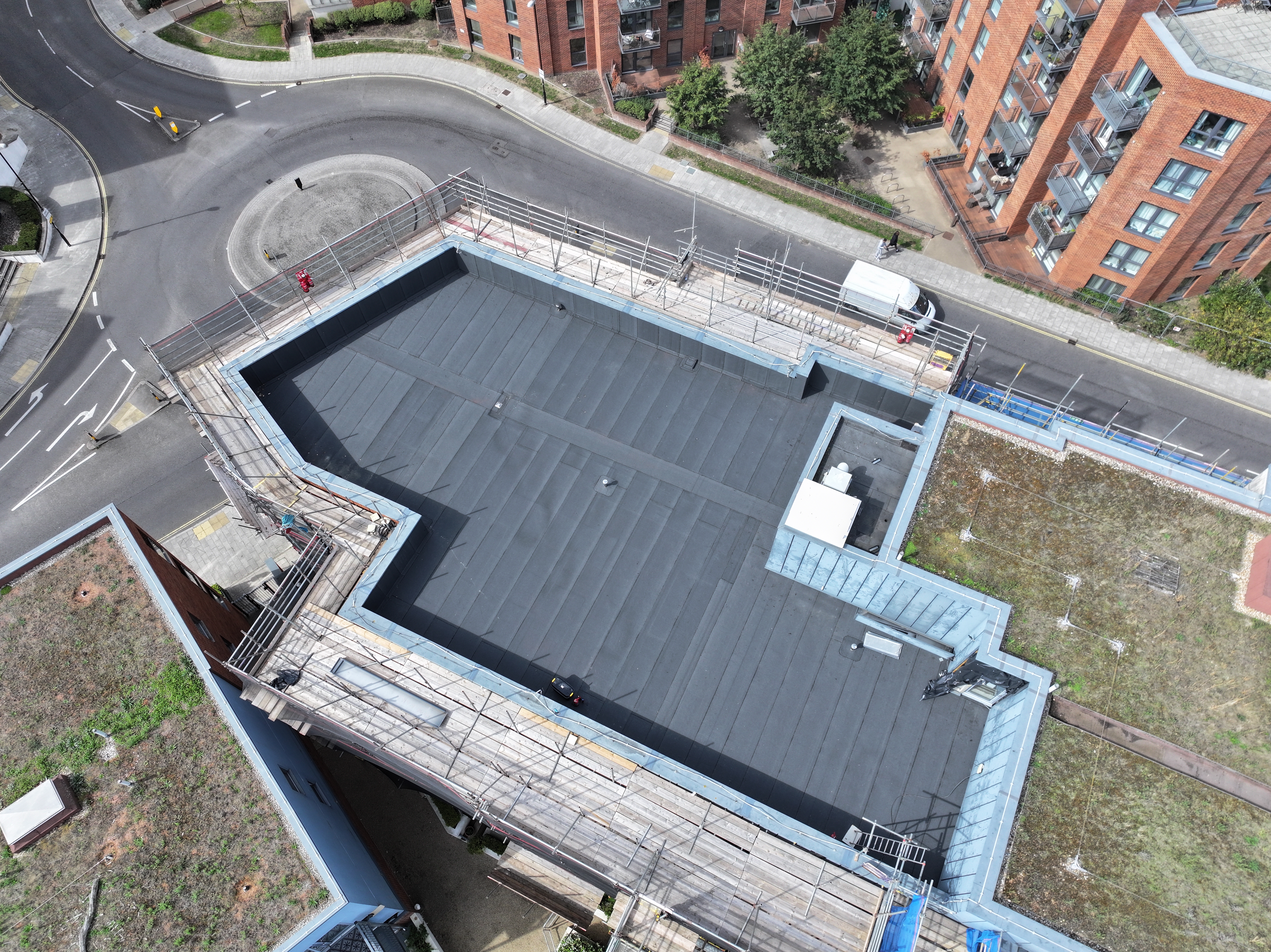 Aerial view of modern building with dark grey roof, surrounded by curved road, red brick residential buildings, and green spaces.
