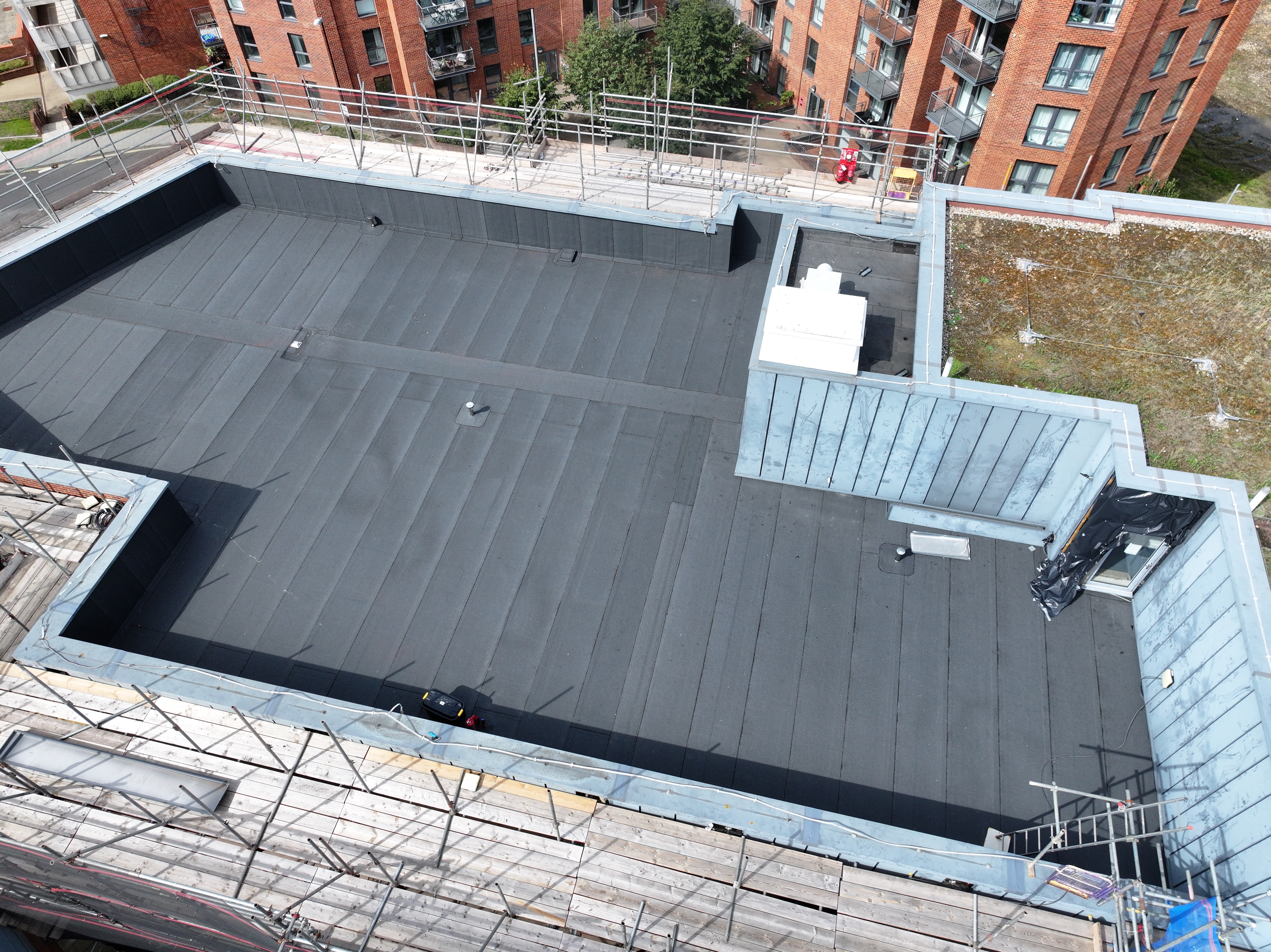 Aerial view of flat rooftop with dark grey membrane, scaffolding around edges, and red brick buildings in background.
