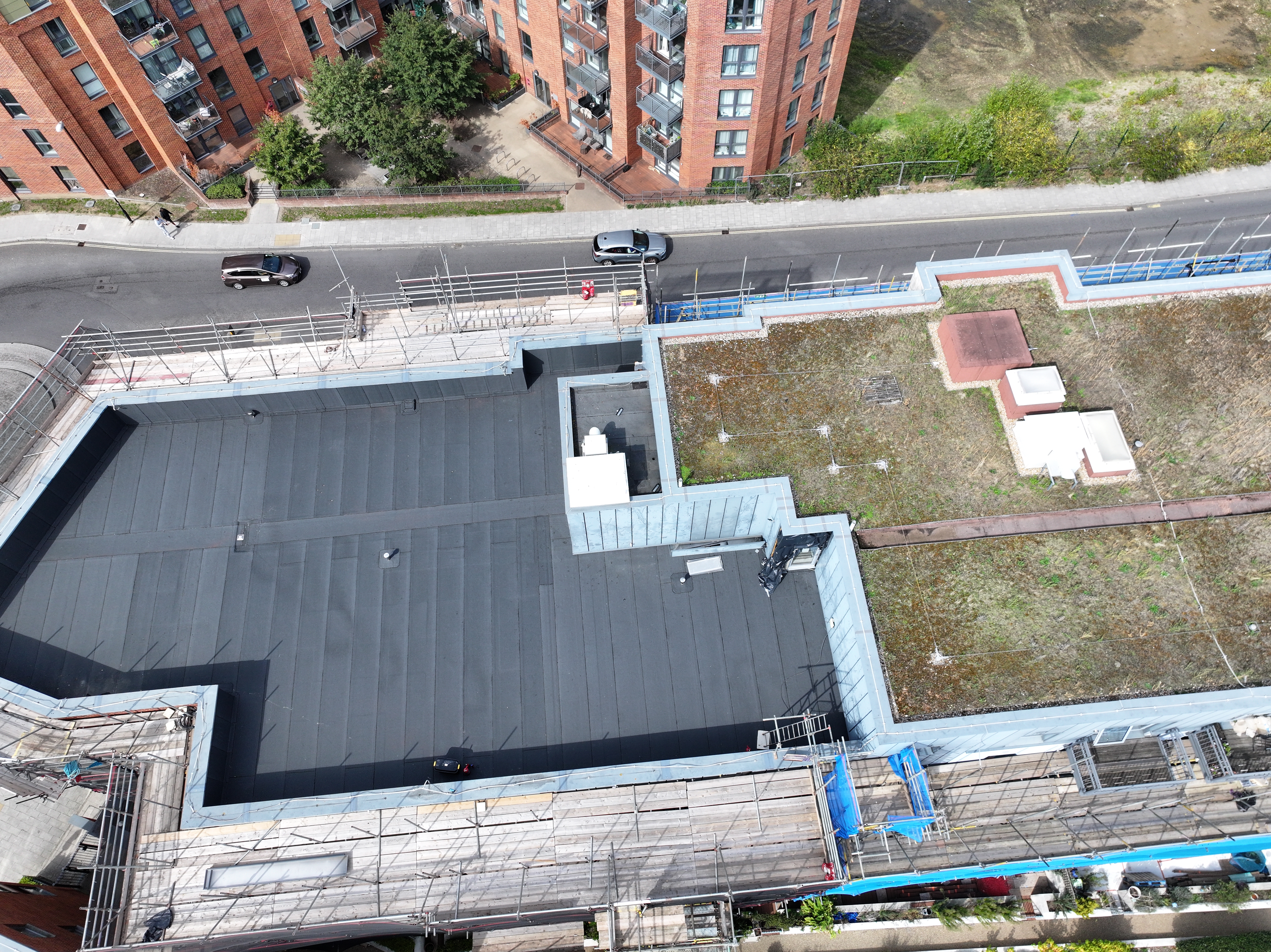 Aerial view of construction site with dark grey roof, white concrete structures, red brick buildings, and surrounding streets with vehicles.