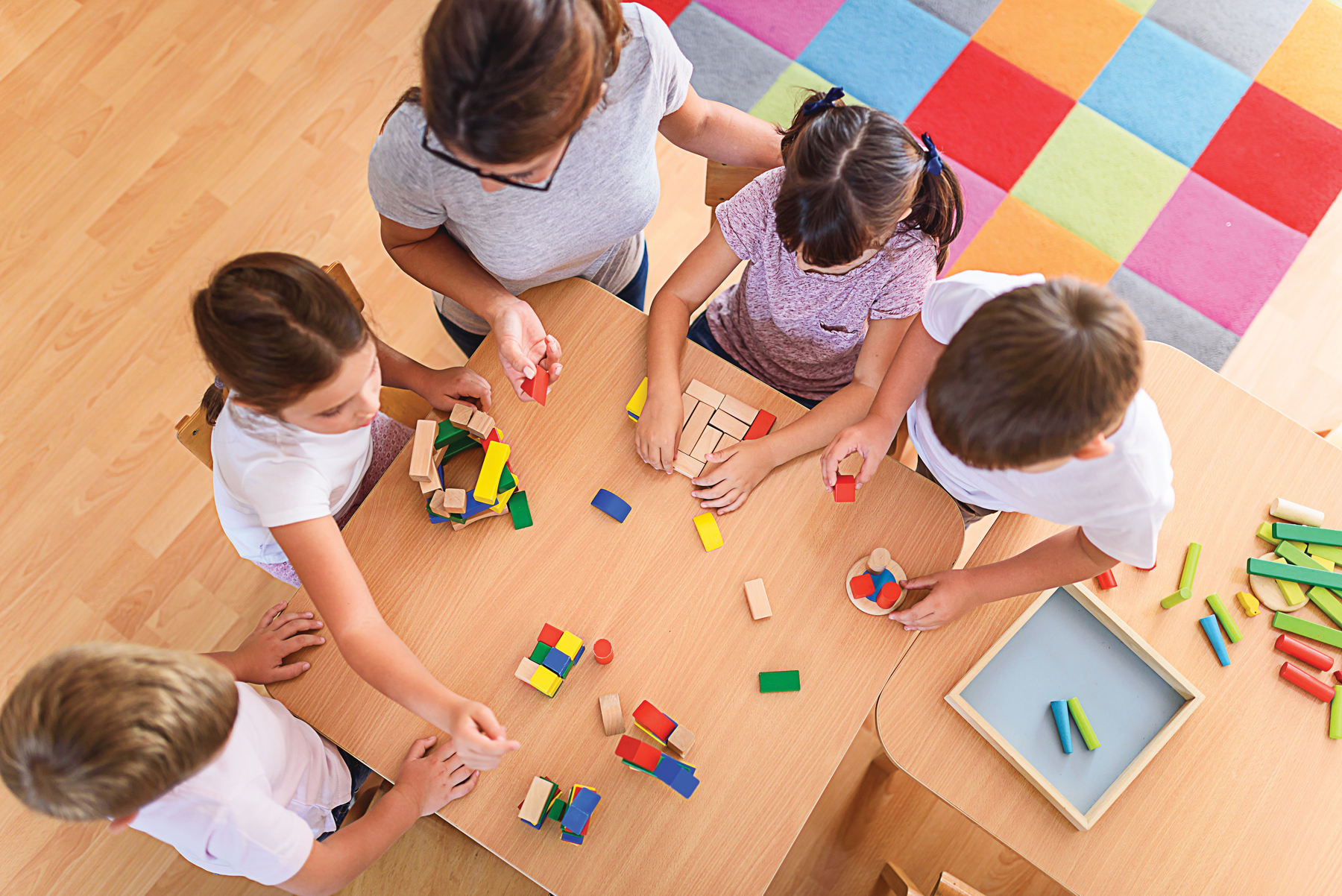 Adult and four children in white tops sitting on wooden floor around coloured building blocks, with multicoloured square carpet behind.