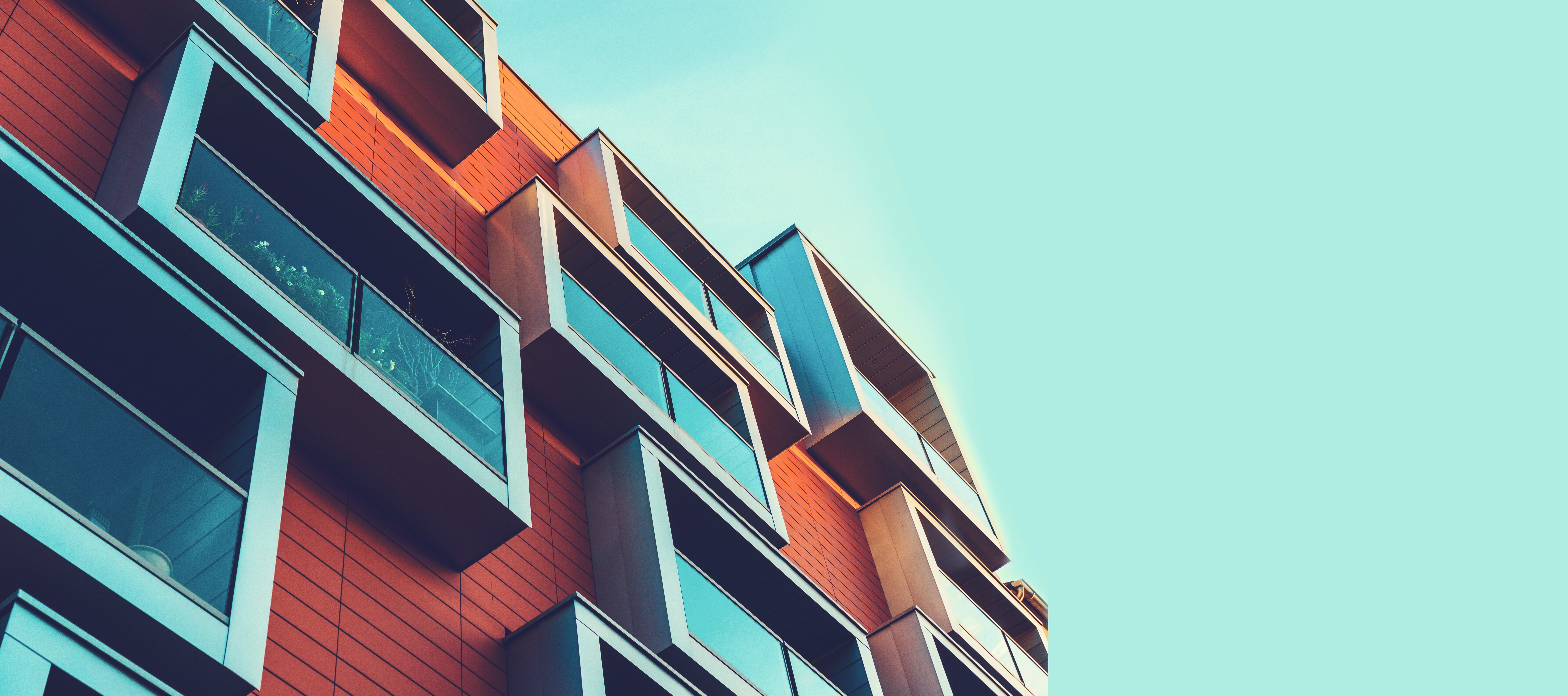 Modern apartment building with orange brick facade and glass balconies shot from below against pale blue sky.