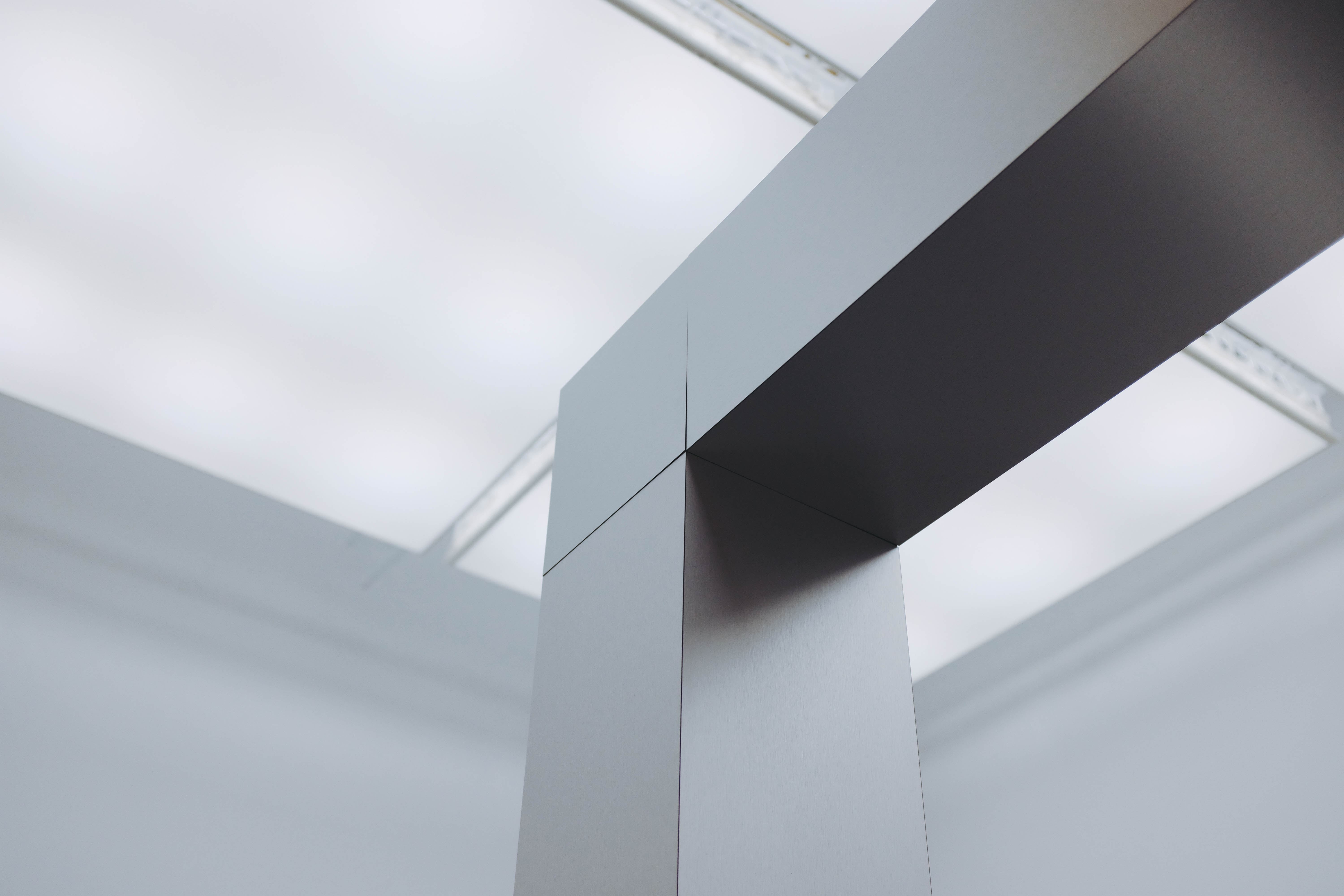 Abstract architectural interior with white geometric walls and dark ceiling beam, featuring angular skylights casting natural light.