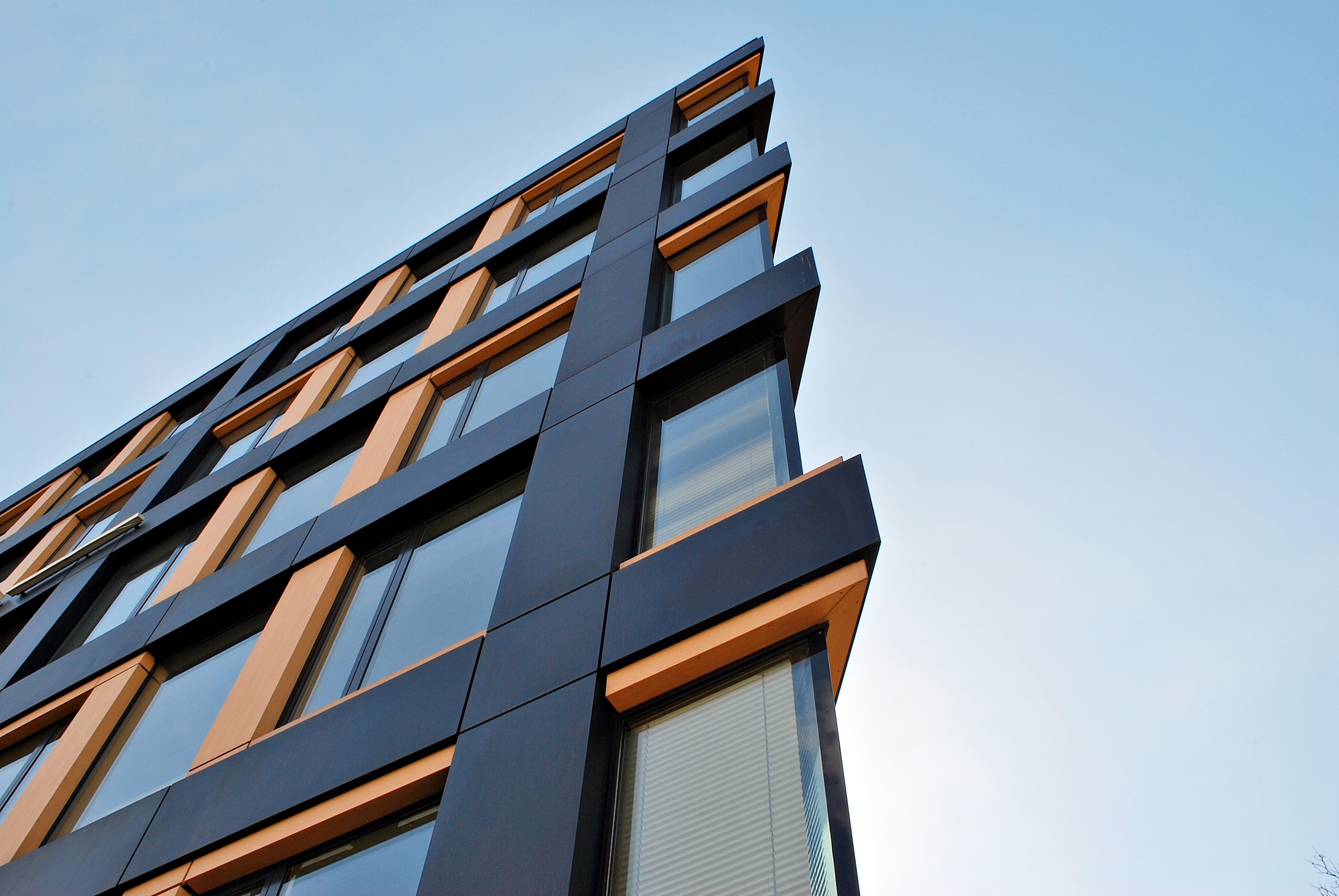 Modern building facade with grey panels, orange window frames, and protruding balconies against blue sky with white clouds.