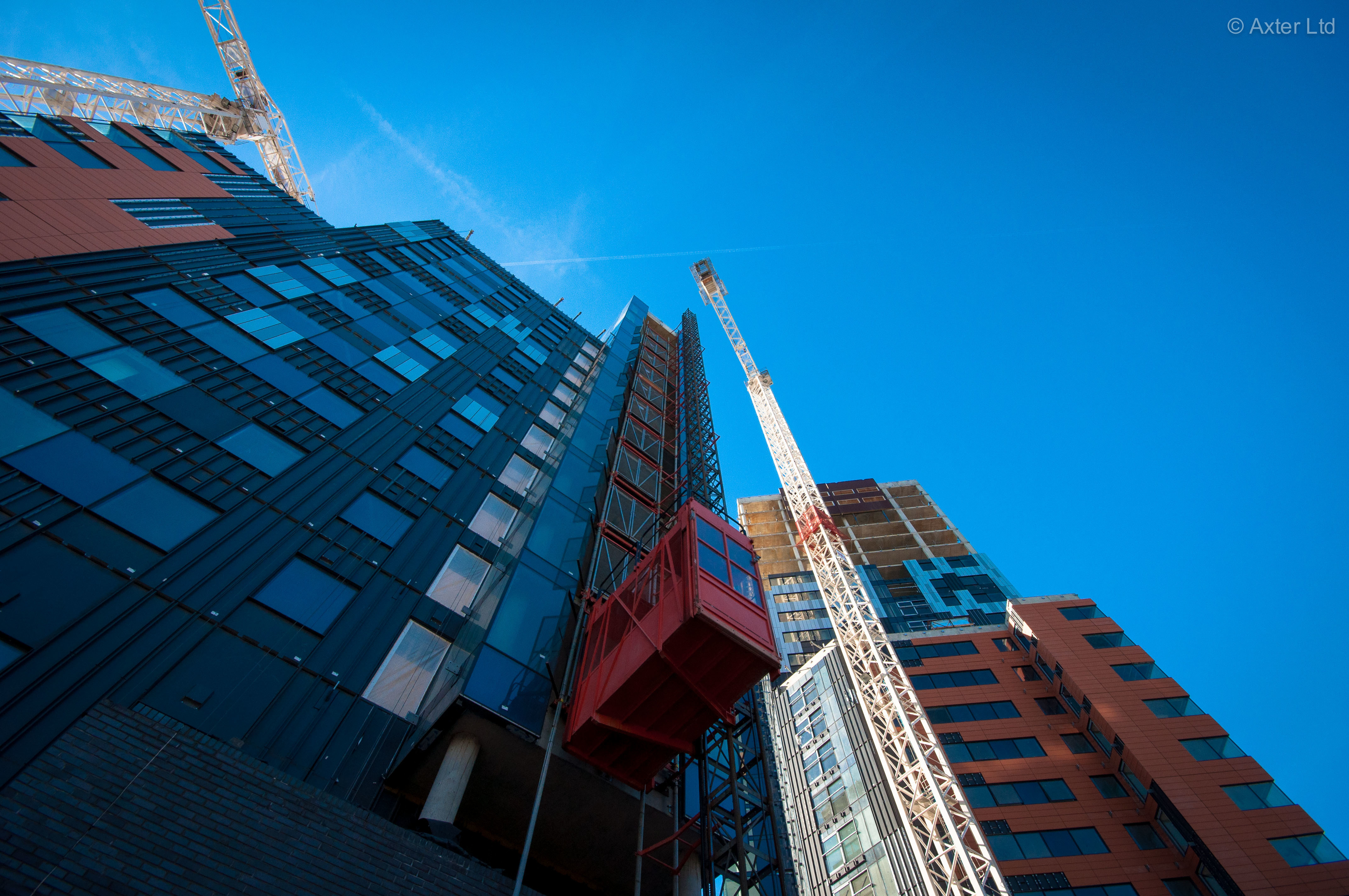 Low-angle view of construction crane extending upward between tall residential buildings with glass facades against bright blue sky.