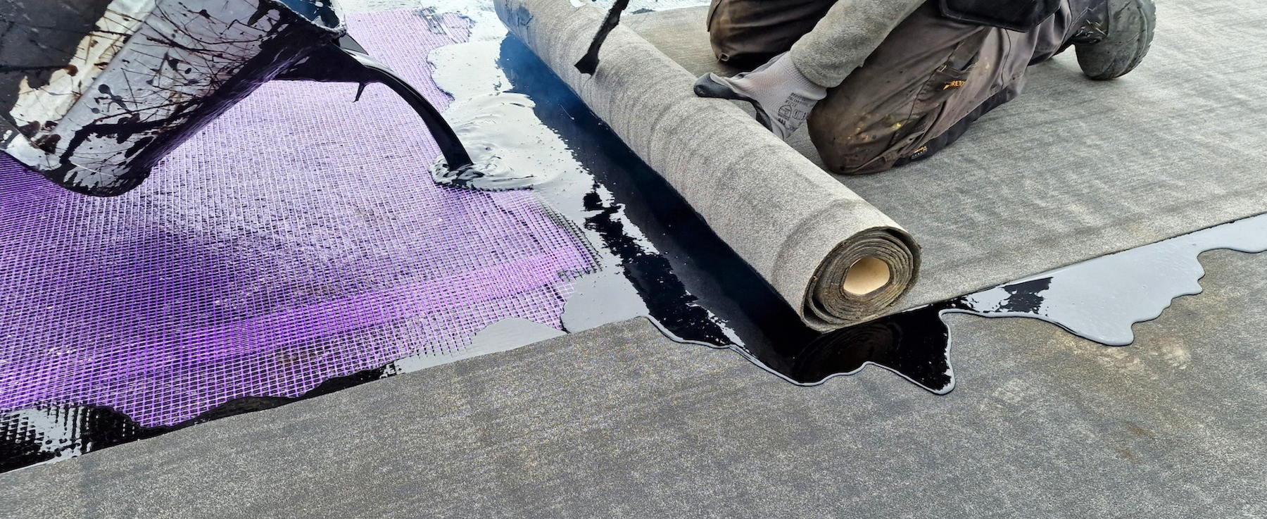 Worker applying black adhesive to concrete floor with purple underlayment material partially rolled out nearby.