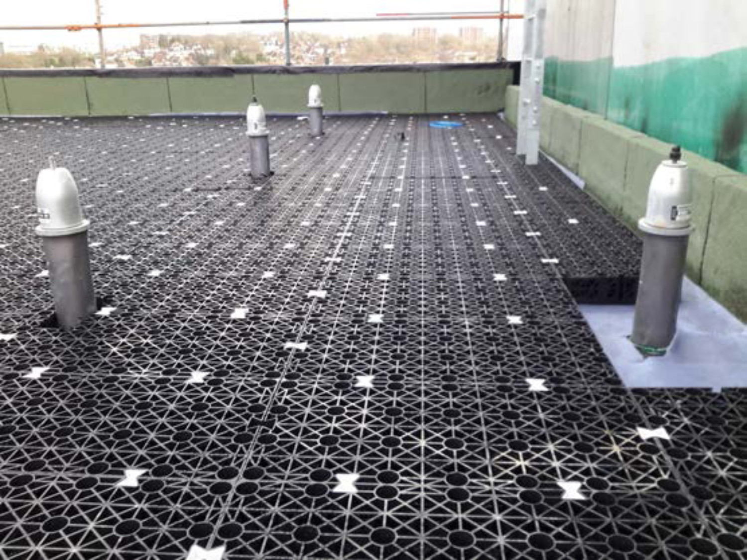 Black geometric grid material covering rooftop surface with cylindrical grey pipes protruding upwards at regular intervals.
