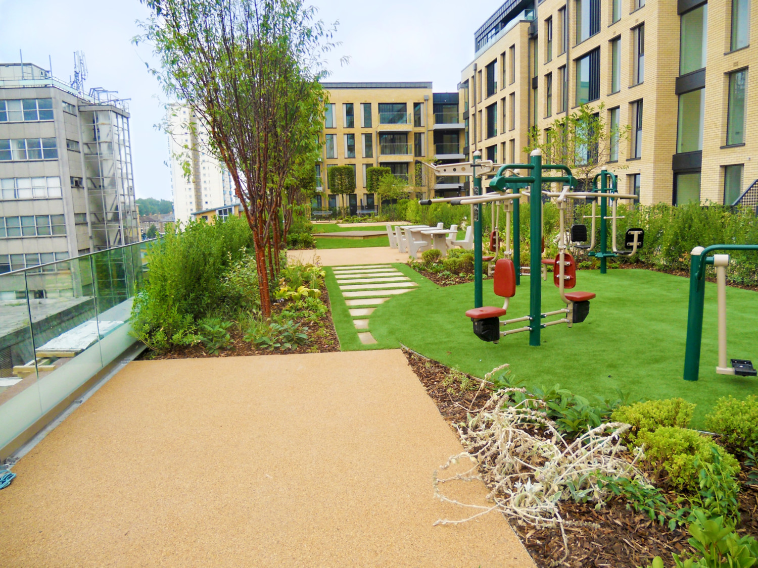 Modern residential courtyard with green exercise equipment, artificial turf, concrete pathways, and landscaped gardens between multi-storey buildings.