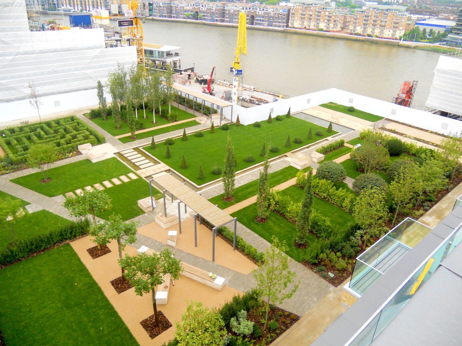 Aerial view of landscaped park with geometric green lawns, pathways, and planted areas beside river with construction cranes visible.