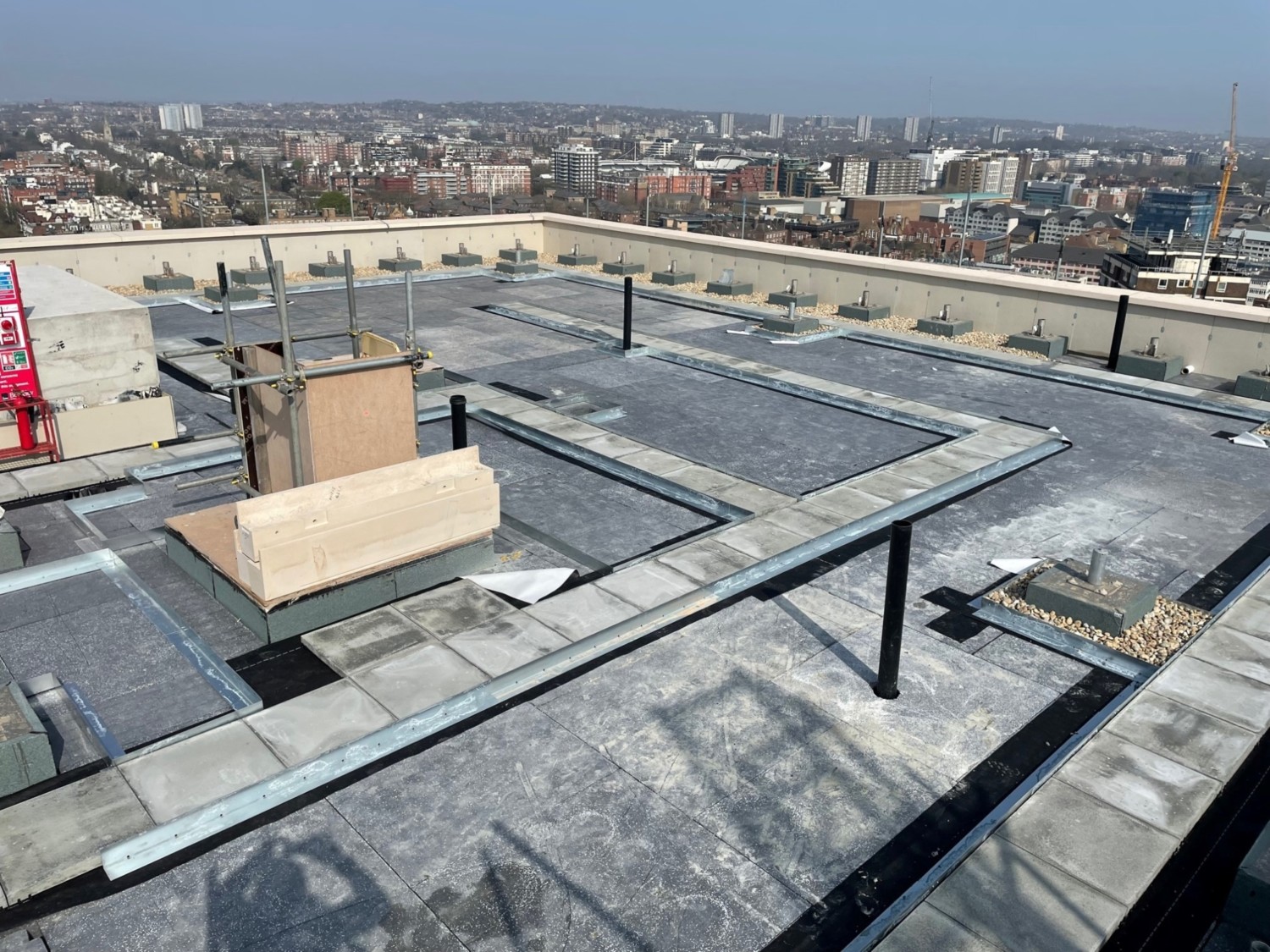 Flat rooftop with grey membrane surface, ventilation units, and metal railings overlooking urban cityscape with buildings and clear sky.
