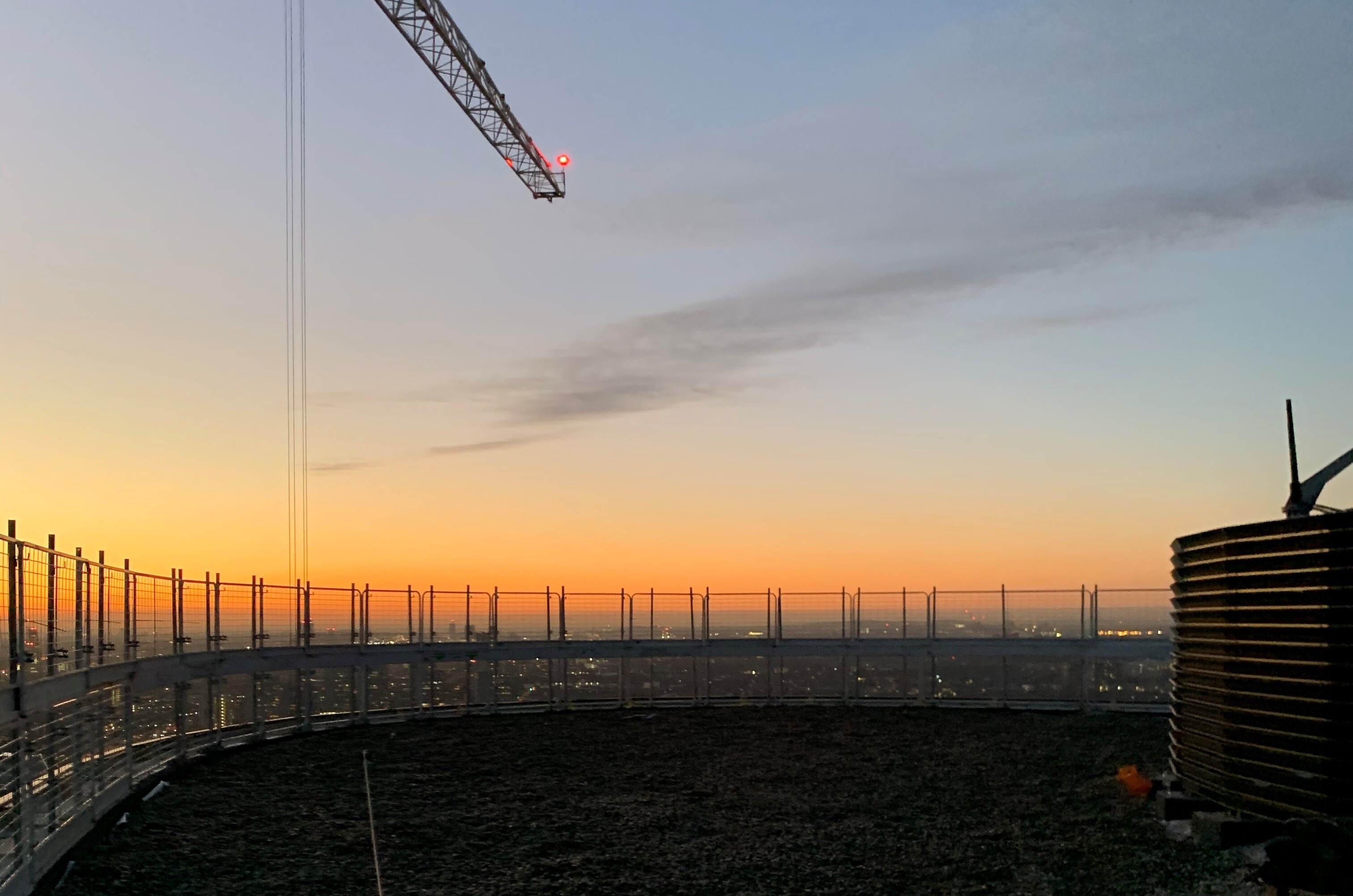 Construction site at dusk with crane overhead, concrete pillars in rows, and orange-pink sunset sky with grey clouds.
