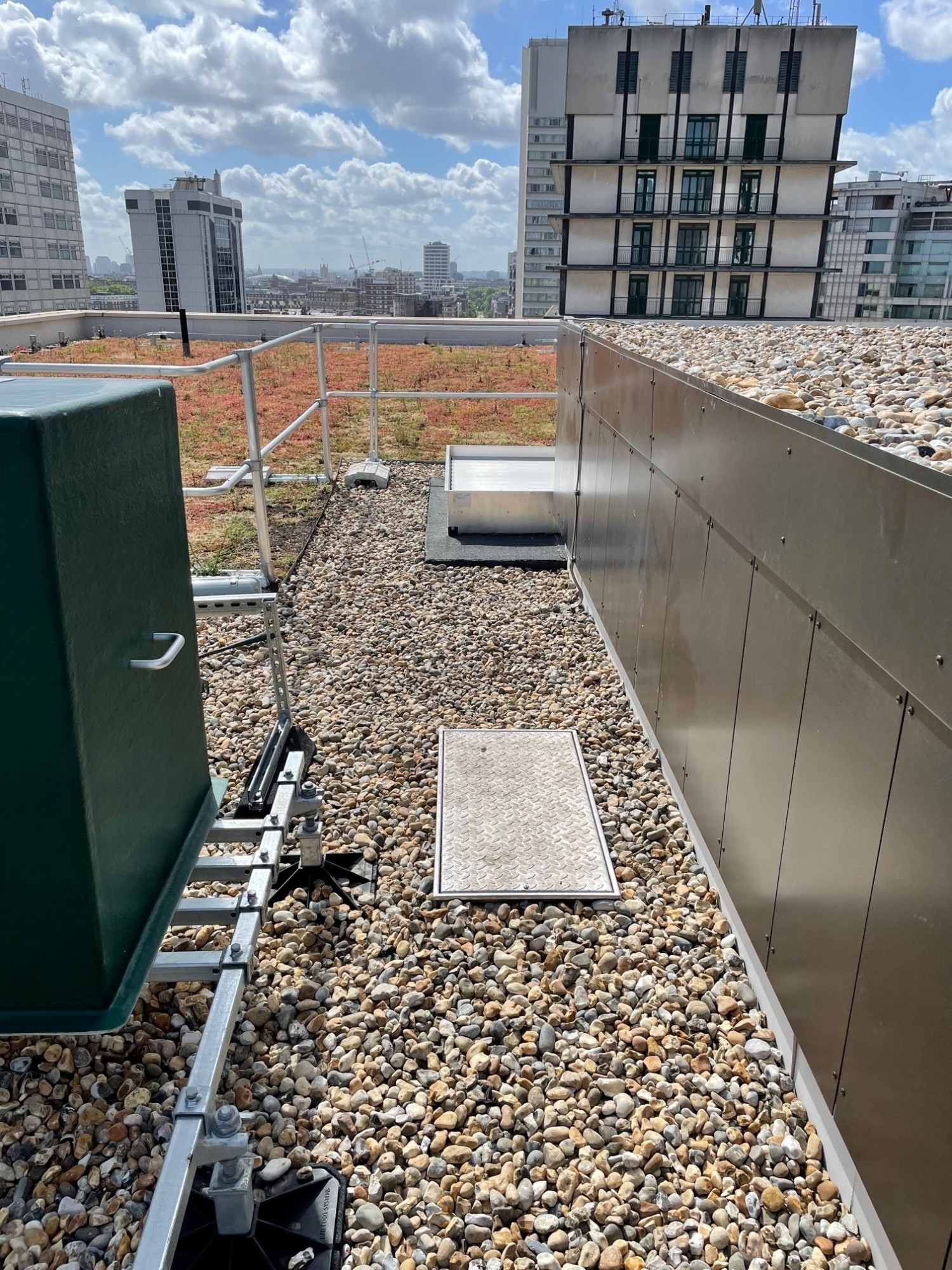 Rooftop with gravel surface, green equipment box, metal ventilation units, and city buildings under cloudy blue sky.
