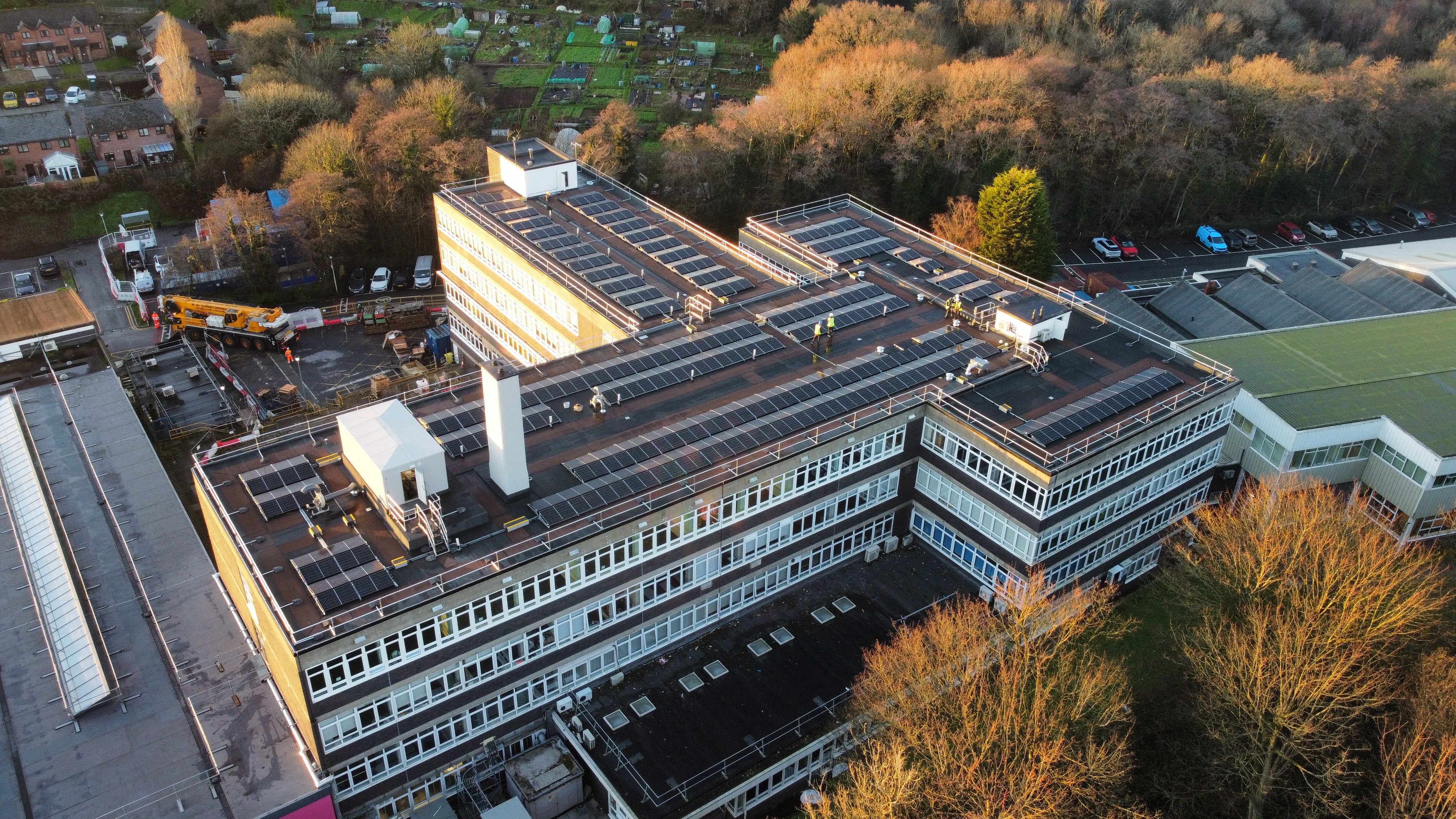 Aerial view of large modern building with solar panels on roof, surrounded by autumn trees with orange and yellow foliage, car parks visible.