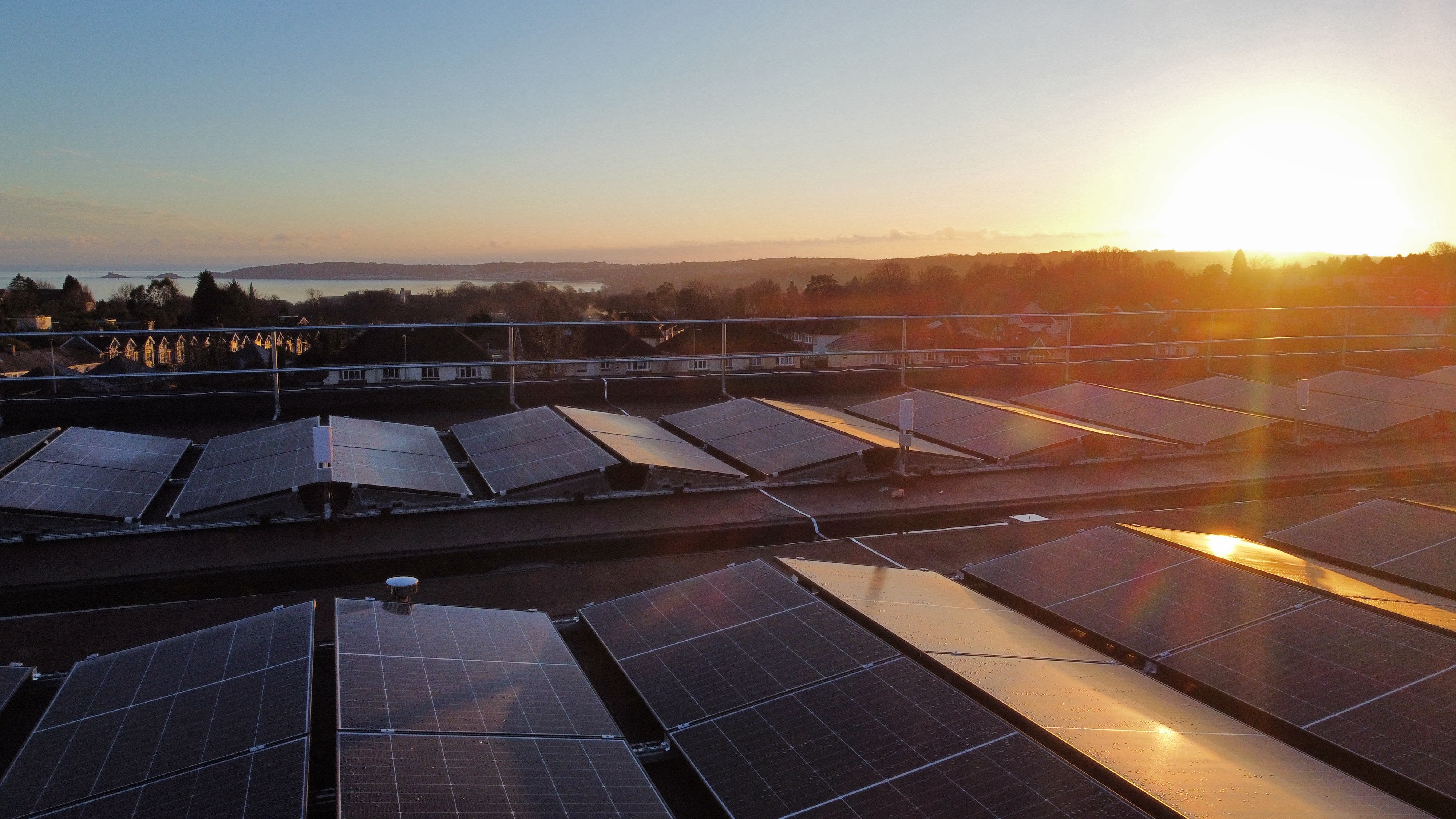 Solar panel array on rooftop at sunset with bright sun flare, residential buildings and countryside in background.