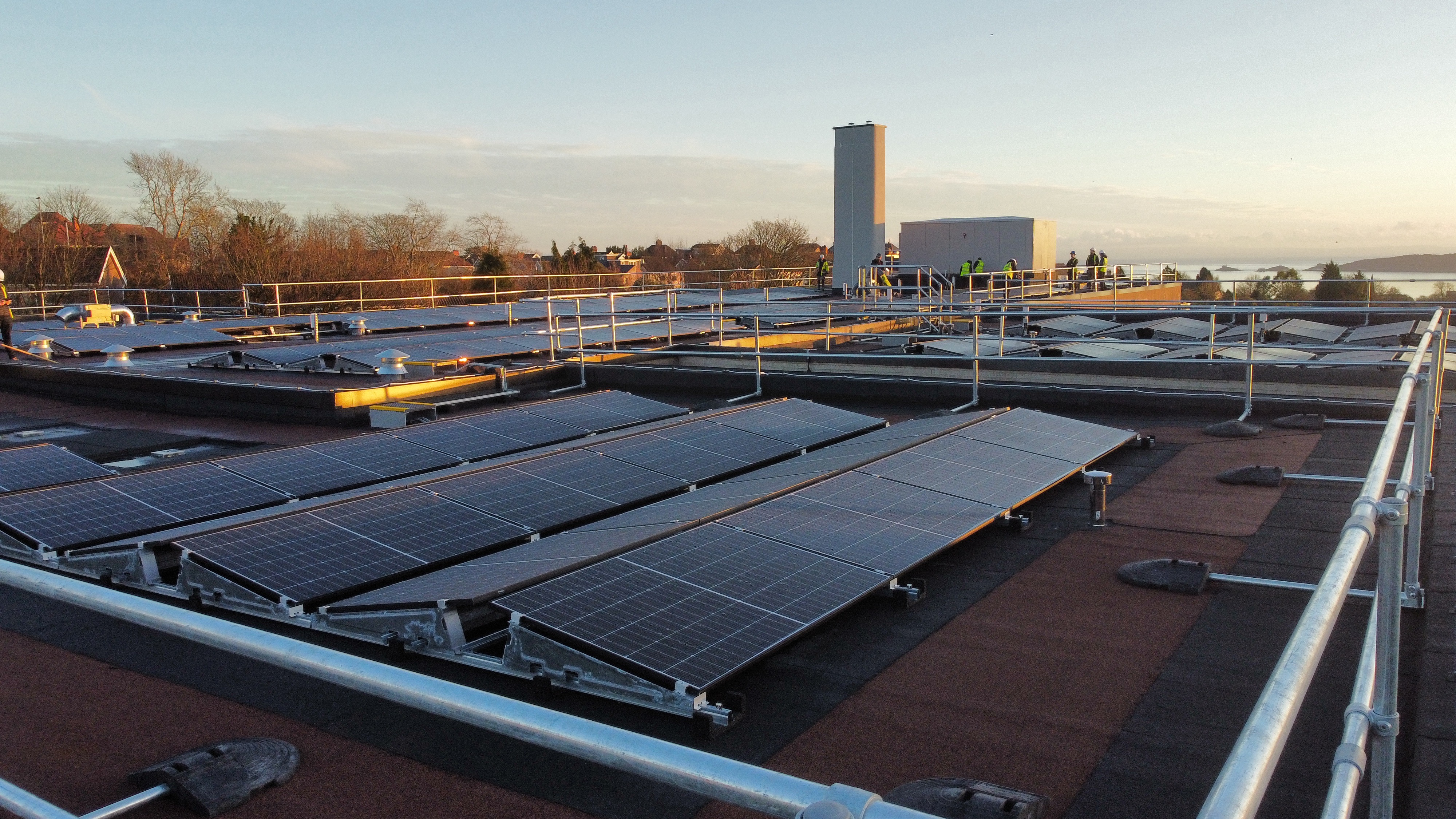 Solar panels installed on flat rooftop with industrial buildings and bare trees visible in background under pale sky.