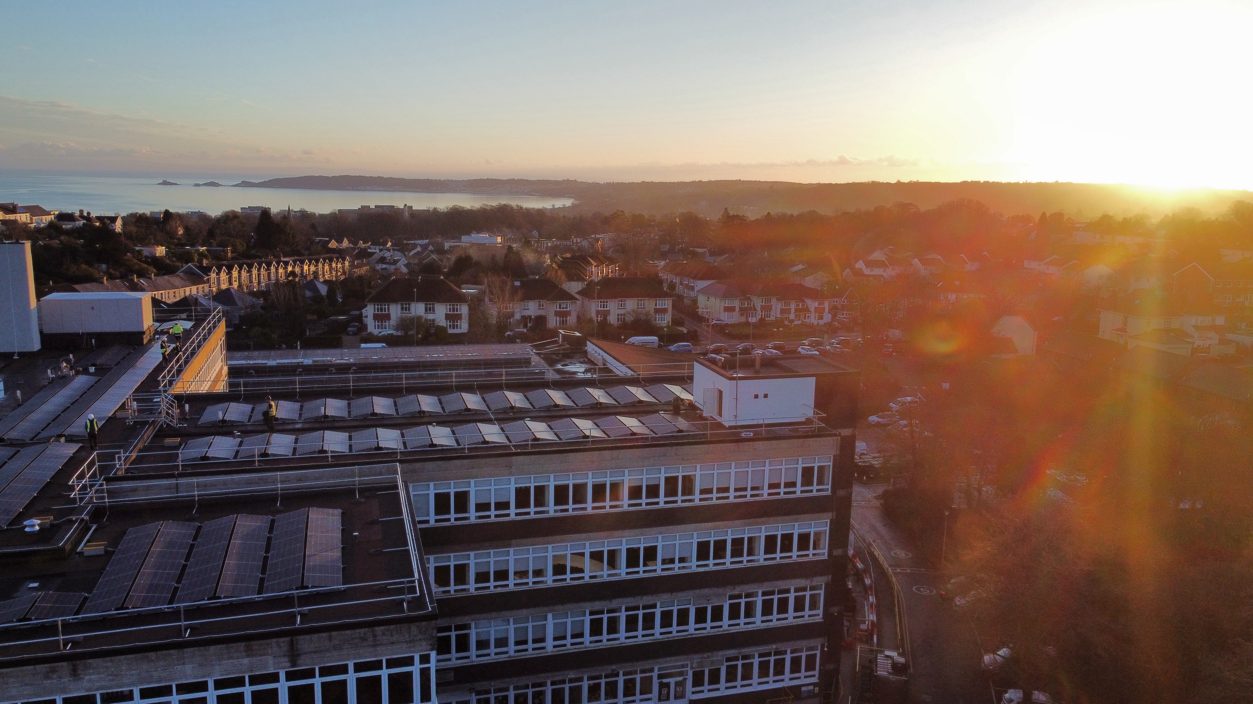 Aerial view of modern buildings with solar panels on rooftops at sunset, with residential areas and hills in background.