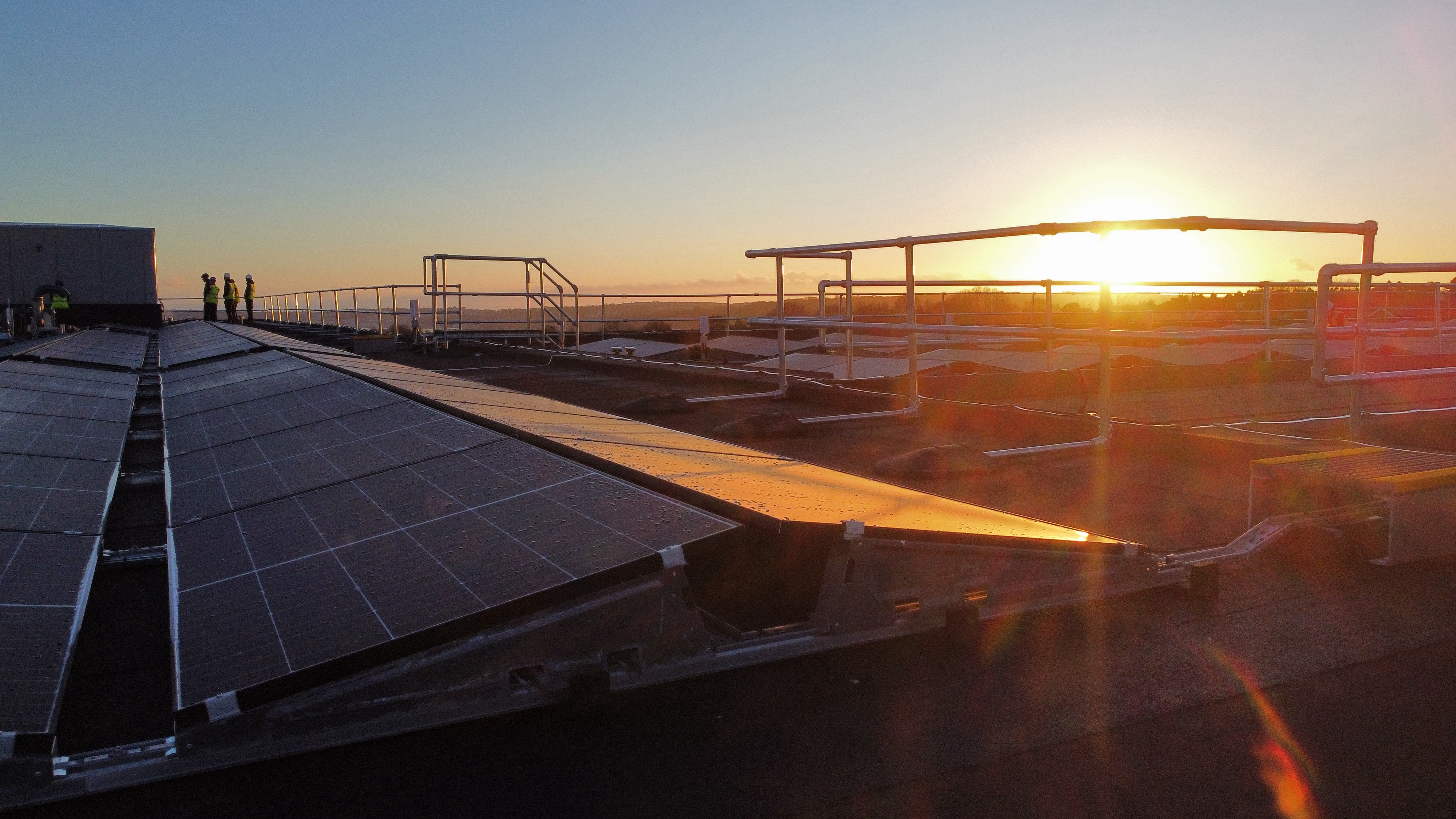 Solar panel installation on rooftop at sunset with bright sun creating lens flare, metal framework visible against orange and blue sky.